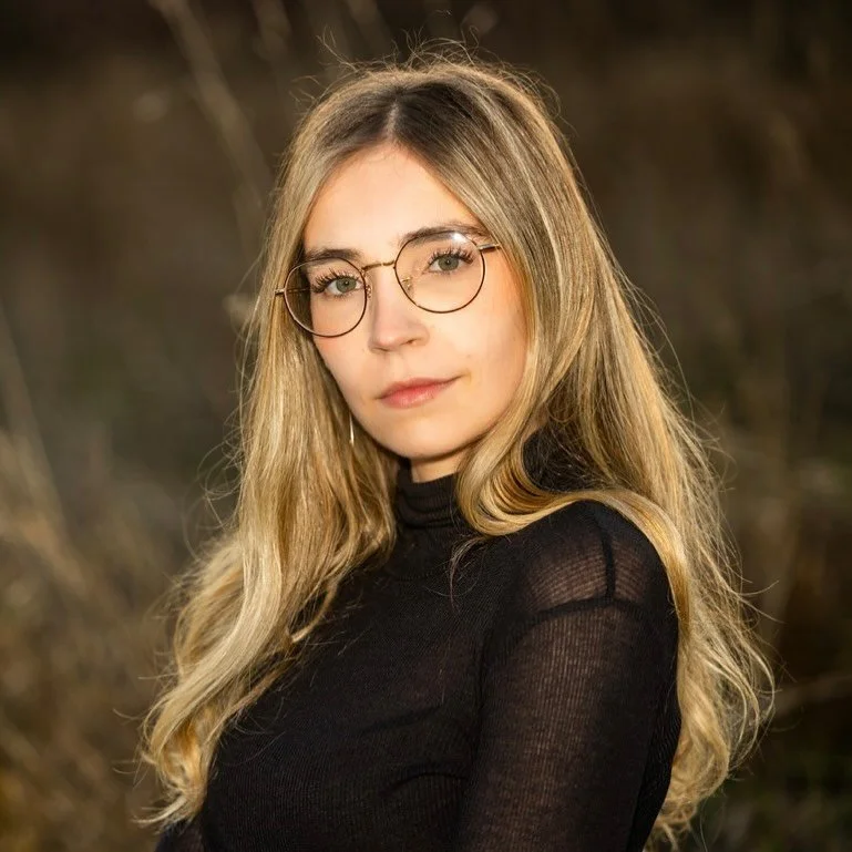 A young woman with long blonde hair wearing glasses and a black top, outdoors with blurred natural background.