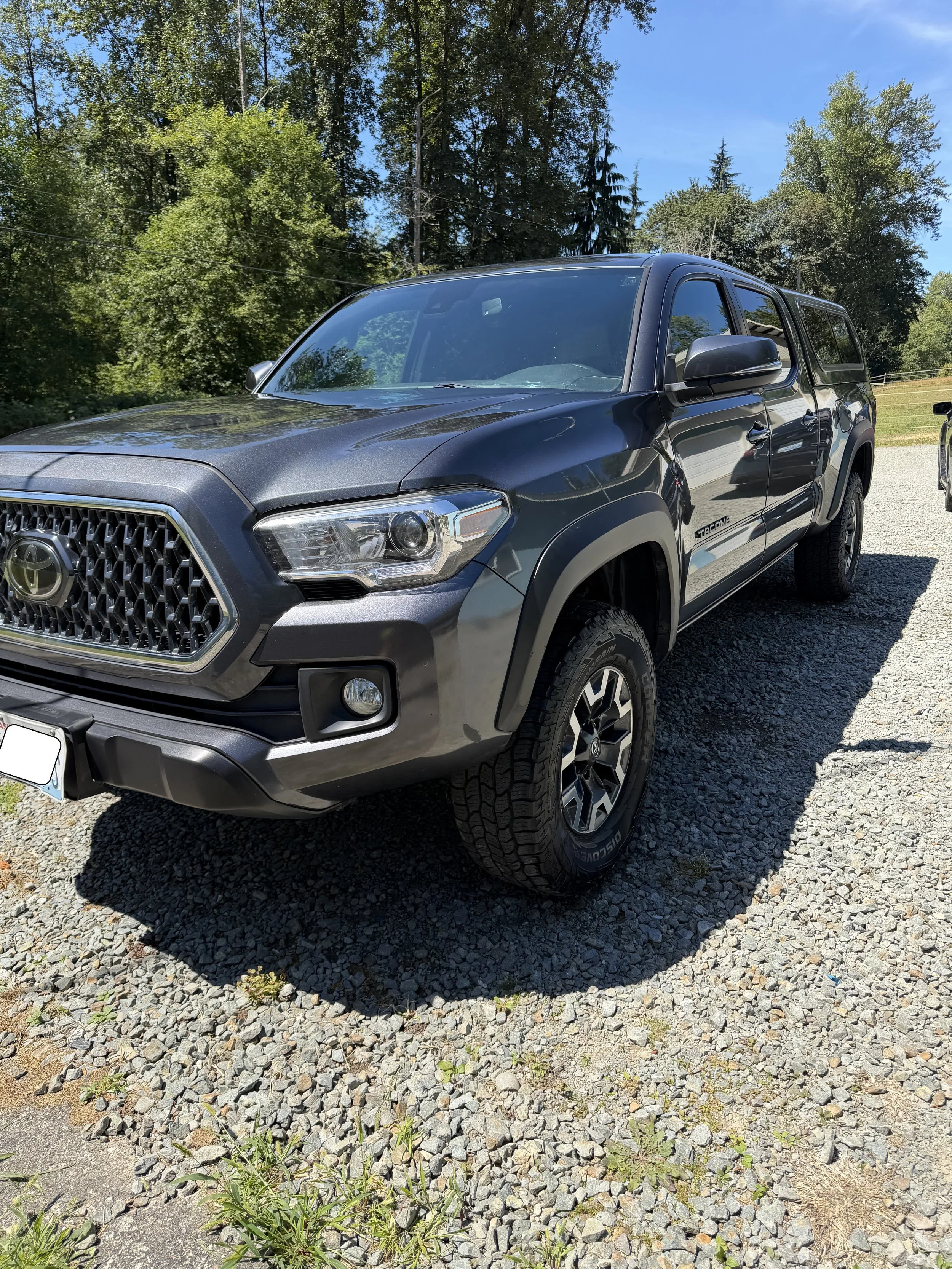 A dark gray Toyota Tacoma pickup truck parked on a gravel surface, with green trees, a grassy field, and a blue sky in the background.