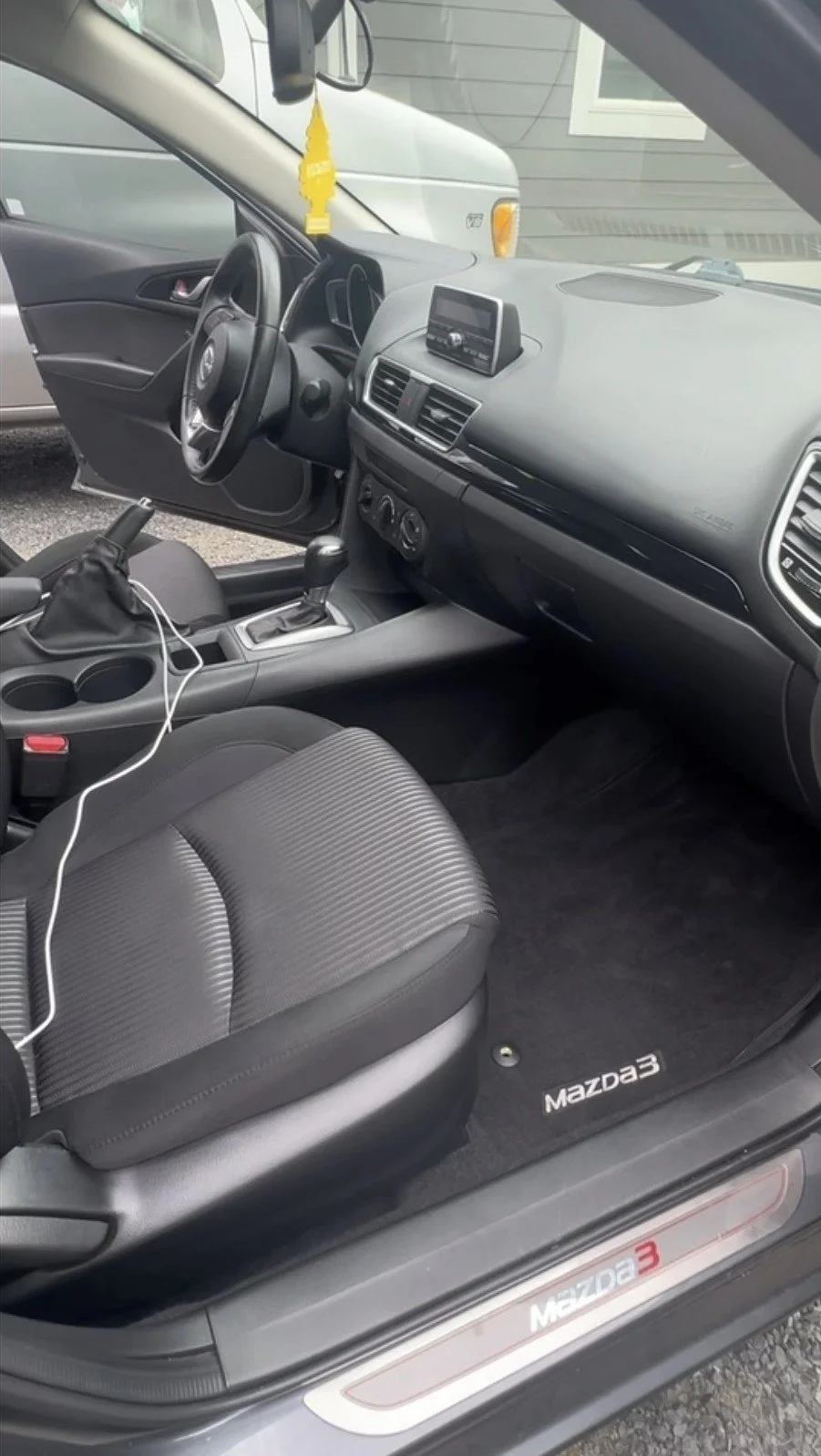 Inside the front cabin of a Mazda 3, showing the driver's seat, steering wheel, dashboard with infotainment screen, gear shift, and center console with cup holders. A white charging cable is connected near the gear shift. The car's floor mat has the Mazda 3 logo.