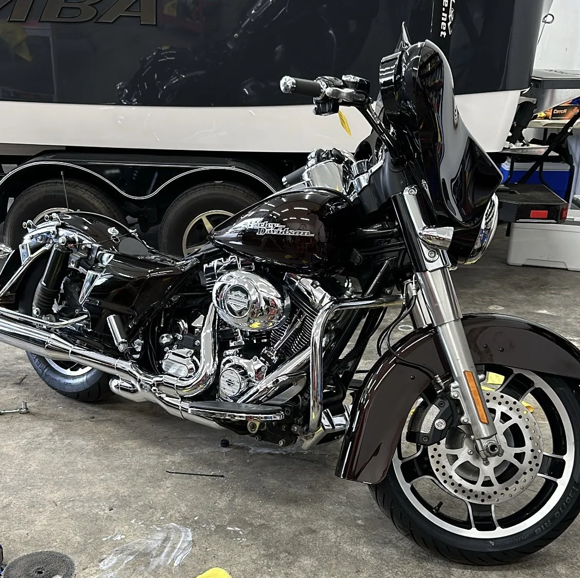 A black Harley Davidson motorcycle parked on a concrete floor in a garage. In the background, there is a large black trailer and some shelves with tools and boxes.