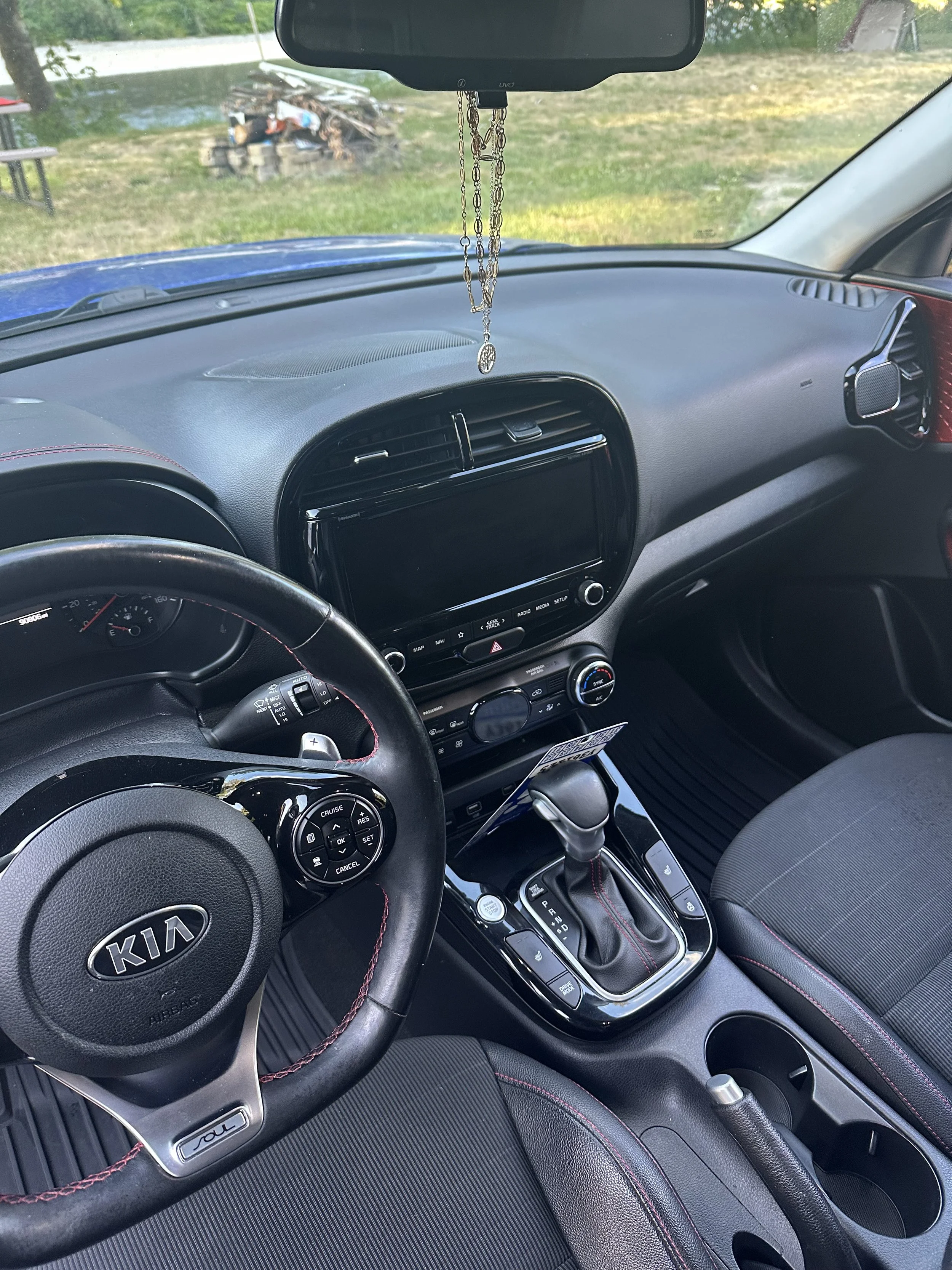 View from the driver's seat of a car showing the dashboard, steering wheel with Kia logo, center console with gear shifter, and hanging rosary beads from the rearview mirror. Outside the windshield, there is a grassy yard with a pile of wood and some outdoor furniture.