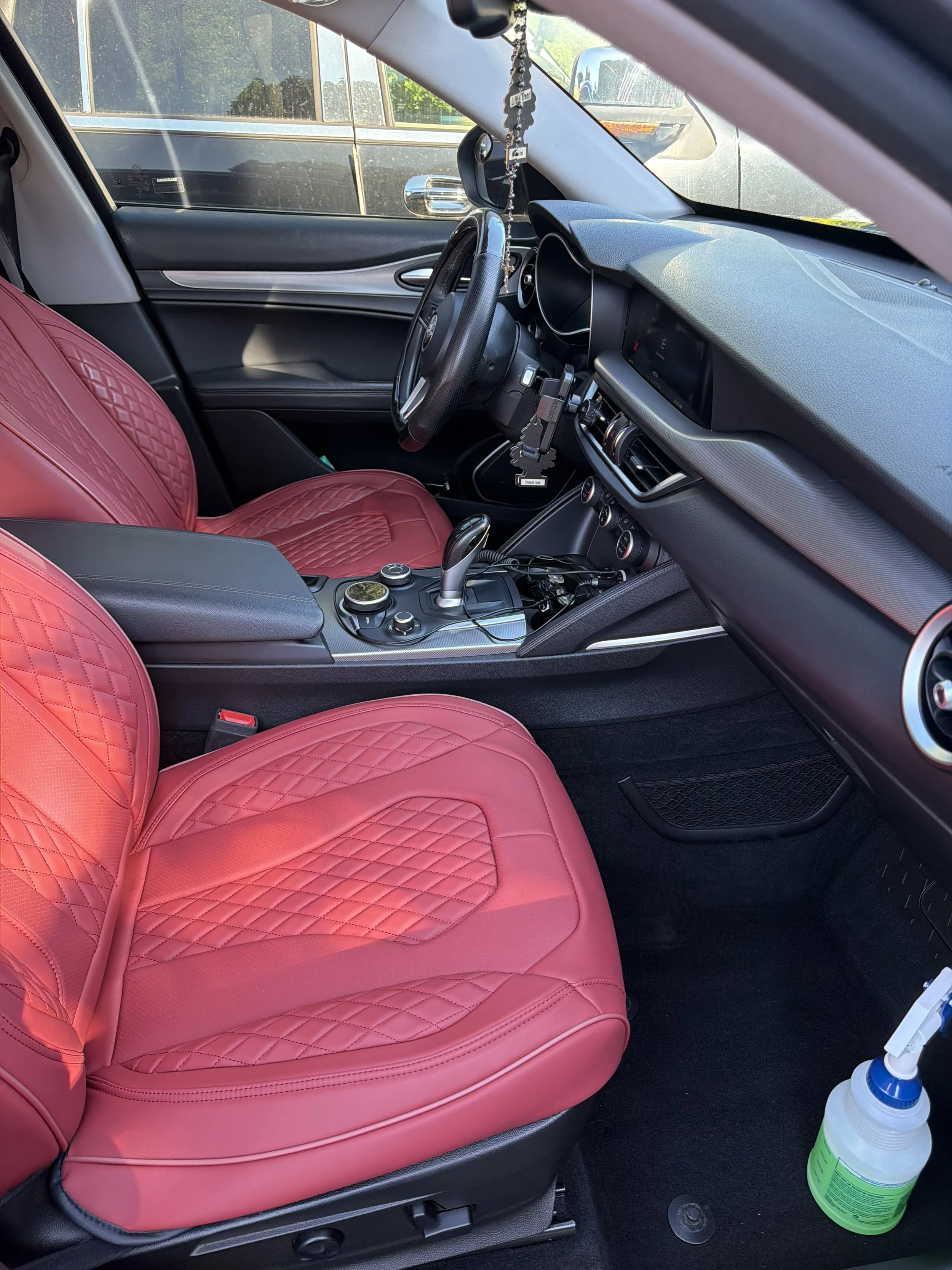 The interior of a car showing the passenger side with a pink quilted seat cover, black dashboard, steering wheel, center console with gear shift, and a bottle of disinfectant on the floor.