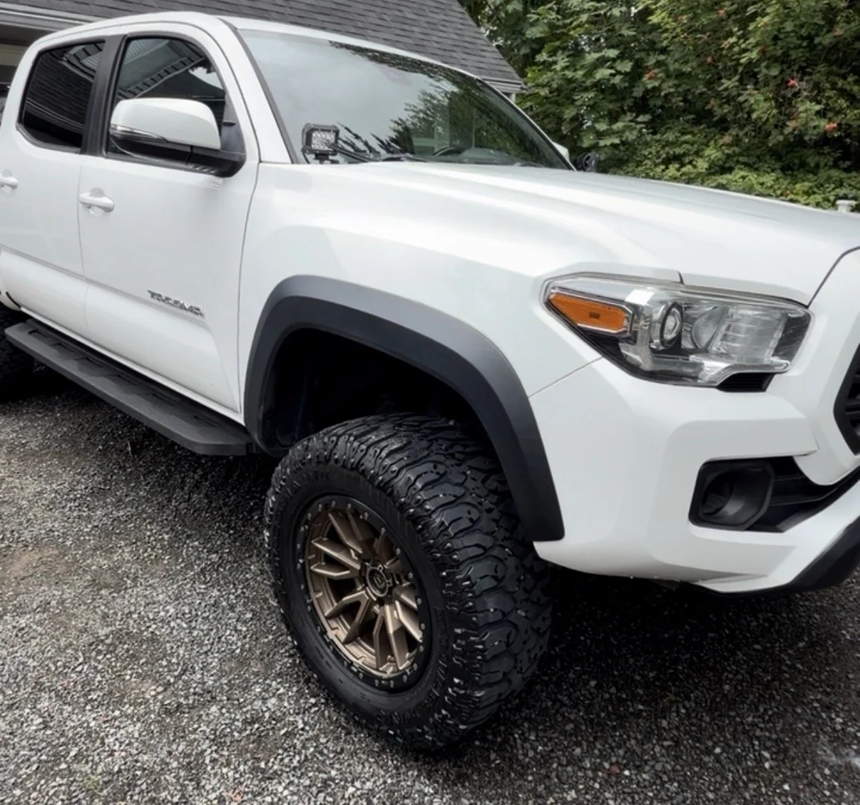 White Toyota Tacoma pickup truck with off-road tires parked on gravel driveway, with green bushes and trees in the background.