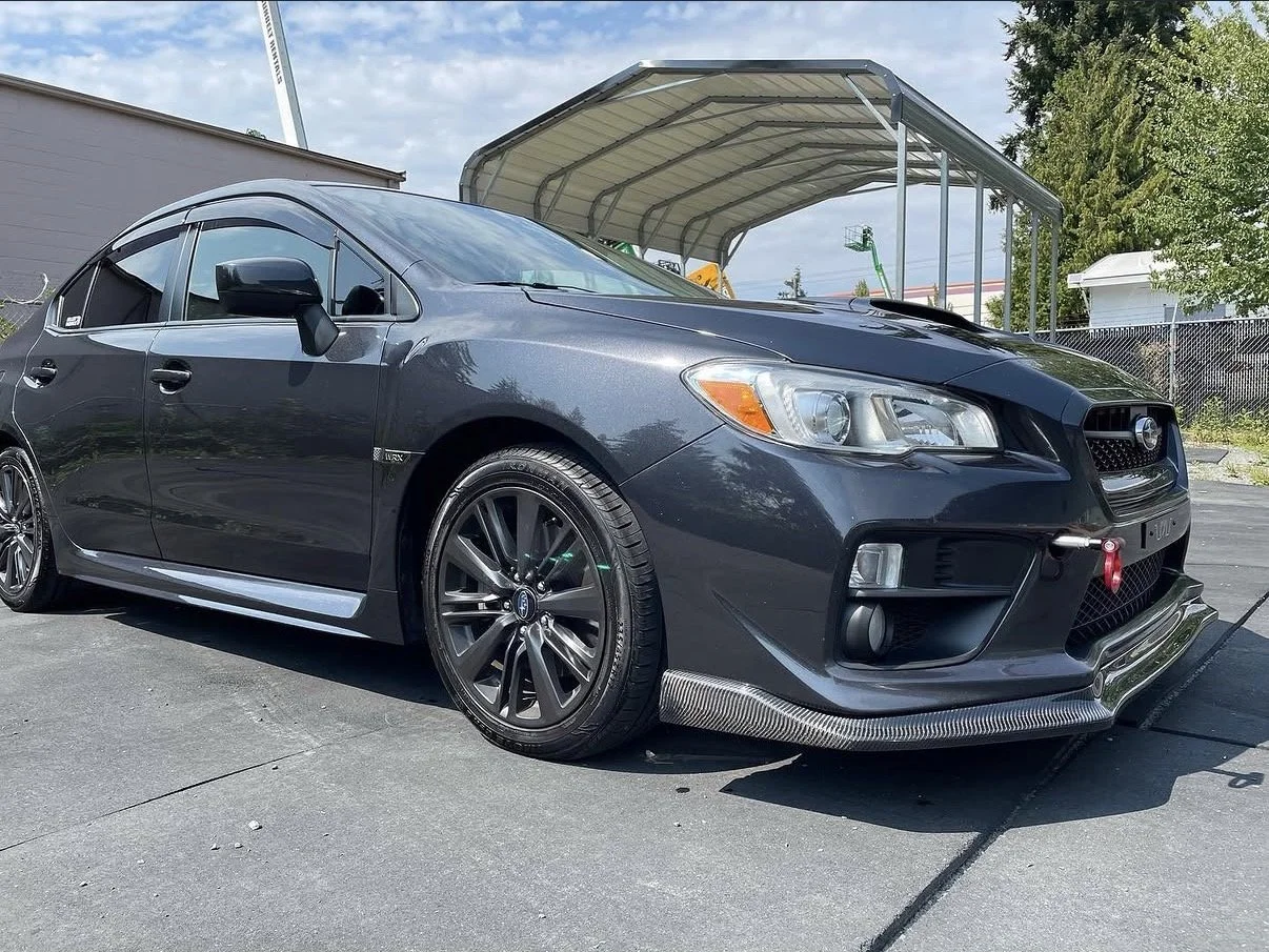 Black Subaru WRX sedan with aftermarket wheels and a carbon fiber front lip, parked outside a carport on a sunny day.