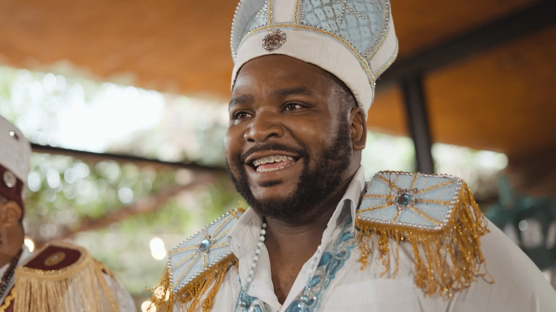 Homem sorrindo usando traje de festa com detalhes em dourado e azul, chapéu decorado com miçangas, em ambiente ao ar livre com fundo desfocado