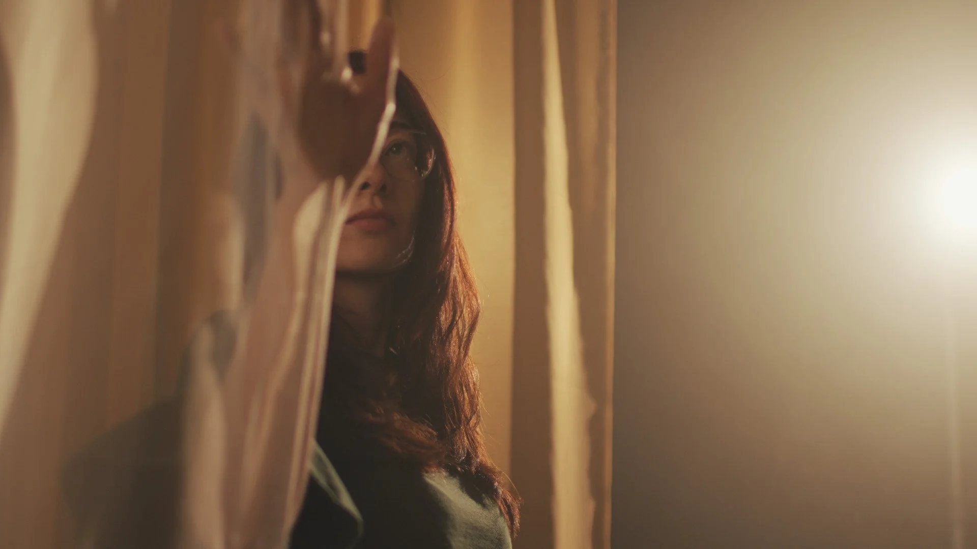 Mulher com cabelo cacheado, vestindo óculos, observando por uma fresta entre cortinas em um ambiente com iluminação quente.