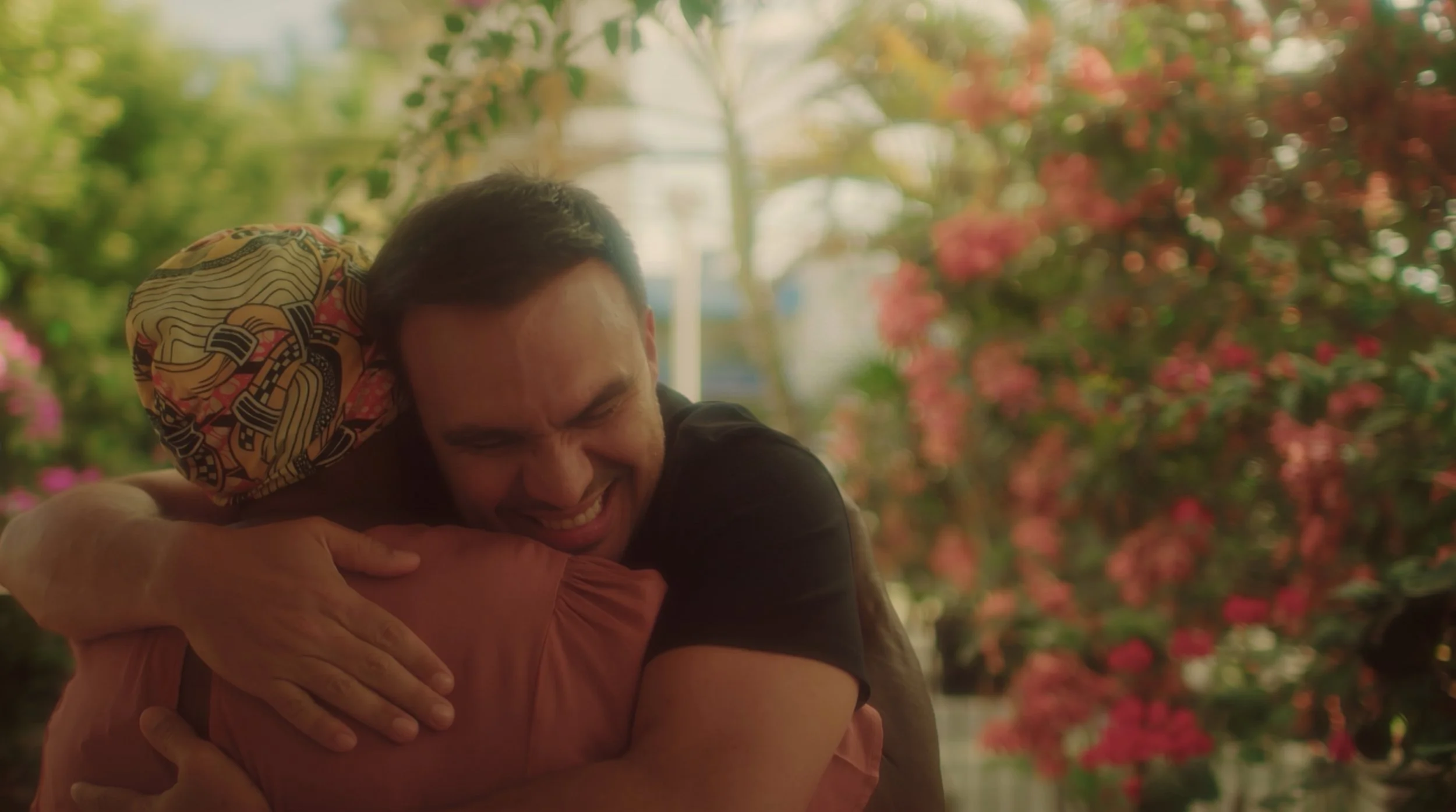 Duas pessoas se abraçando, sorrindo, em um ambiente com muitas plantas e flores ao fundo.
