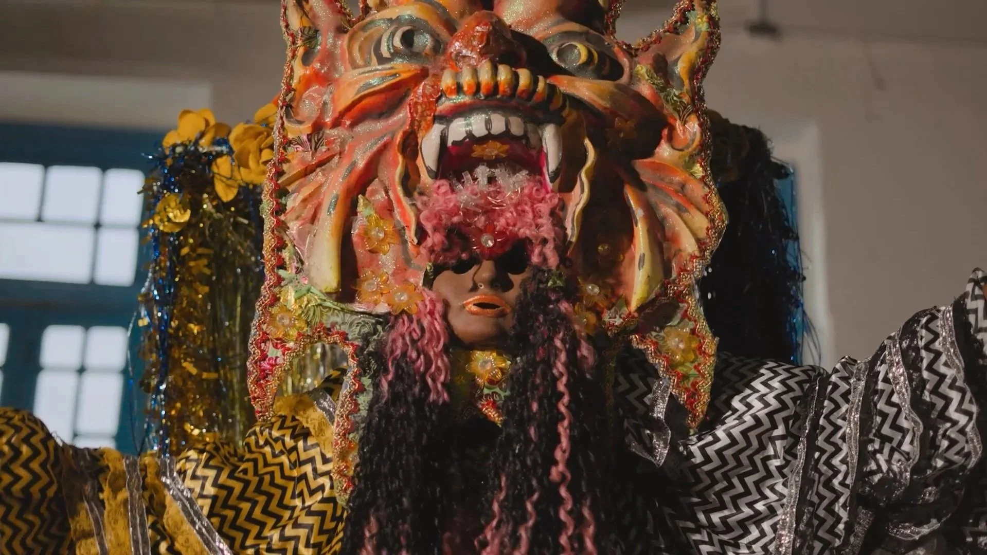 Máscara tradicional de Folia de Reis com detalhes coloridos, um rosto com boca e dentes expostos, como um dragão, e cabelo longo e trançado em cores diversas. A máscara cobre o rosto de uma pessoa com roupas de estampa dourada e preta.