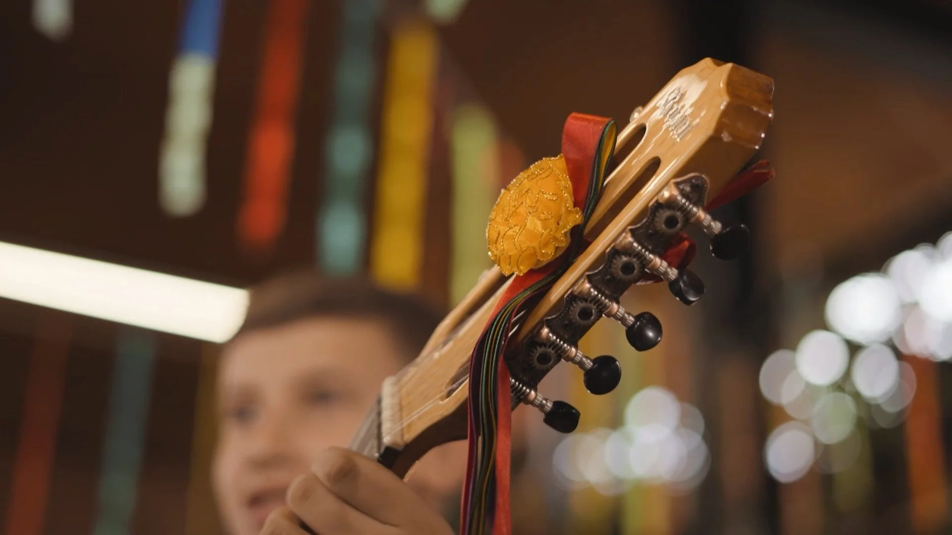 GUITARRA com fio e decoração amarela, sendo tocada por uma pessoa em fundo desfocado com luzes de cores variadas.