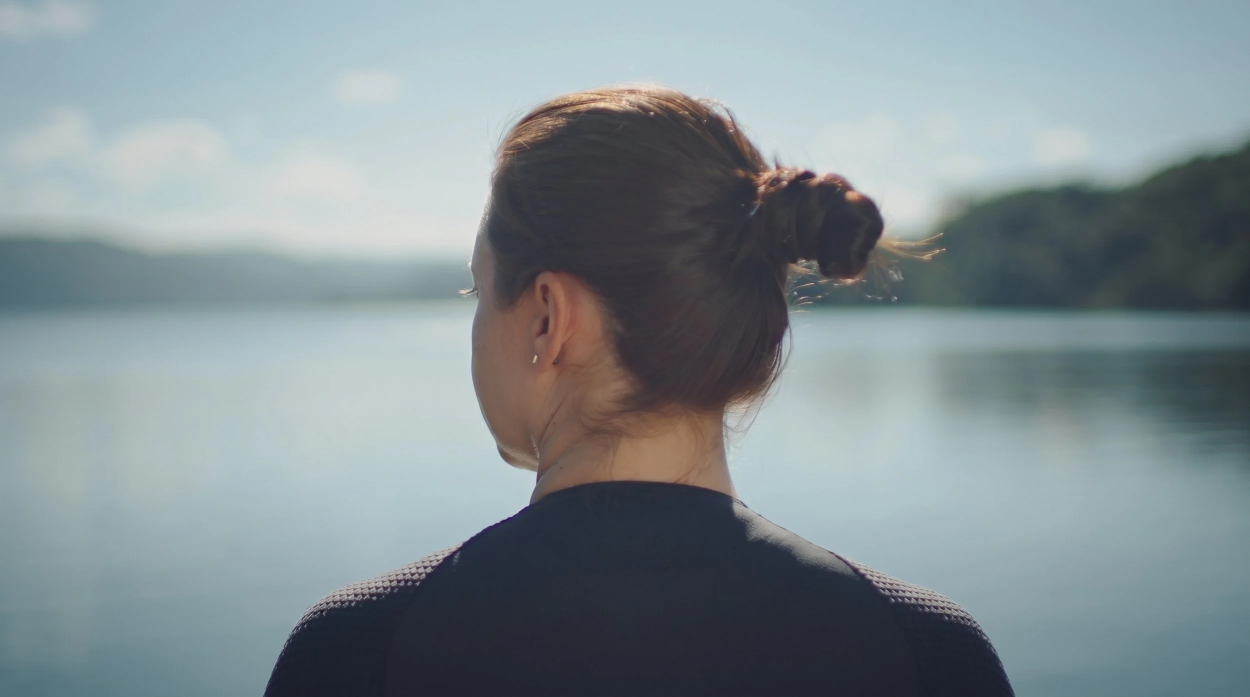 Mulher com cabelo preso em coque, vista de trás, olhando para um rio ou lago sob céu limpo.