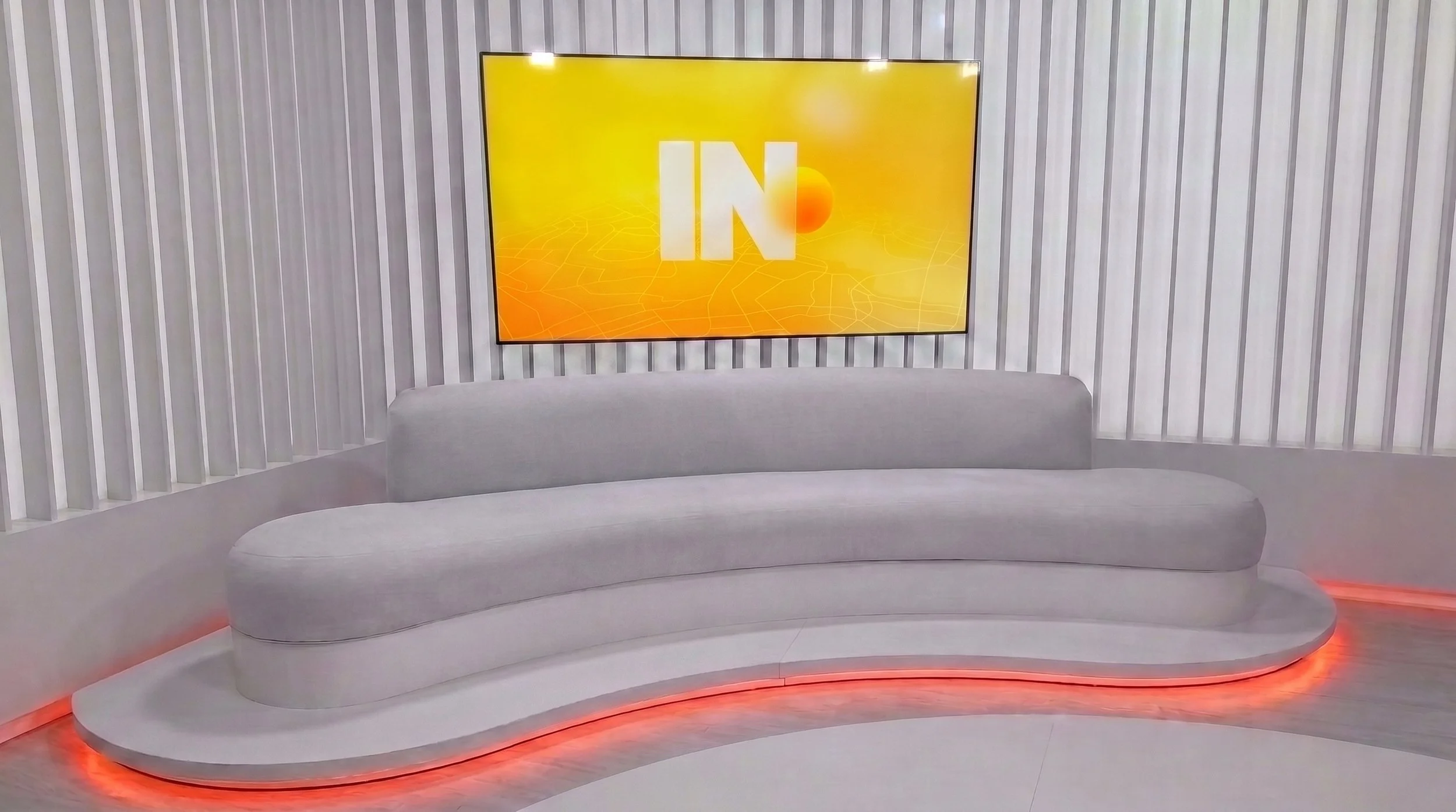 Sala de televisão com sofá cinza e parede com revestimento de madeira e painel de TV empate com fundo amarelo e logo IN na tela.