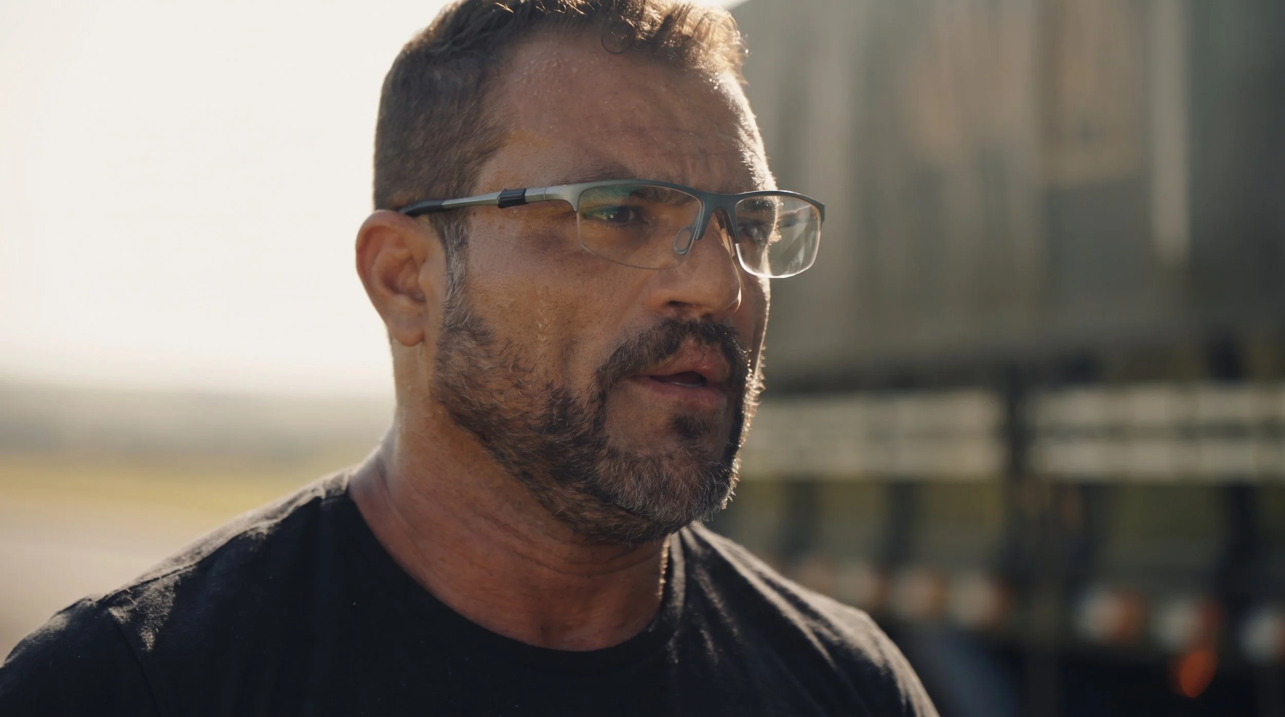 Homem com barba, óculos esportivos e camiseta preta, ao ar livre em dia ensolarado.