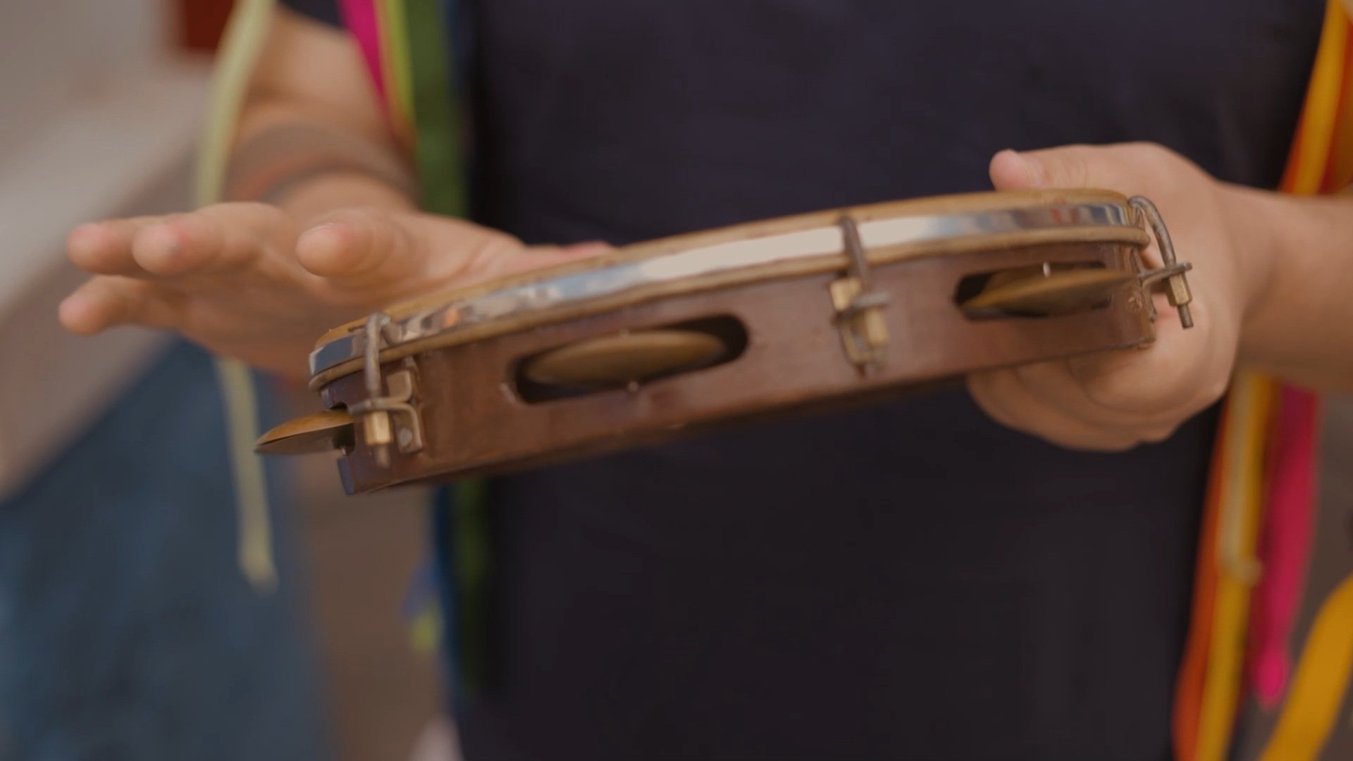 Pessoa segurando um pandeiro de mão, instrumento musical de percussão feito de madeira e metal, com mãos visíveis em foco.