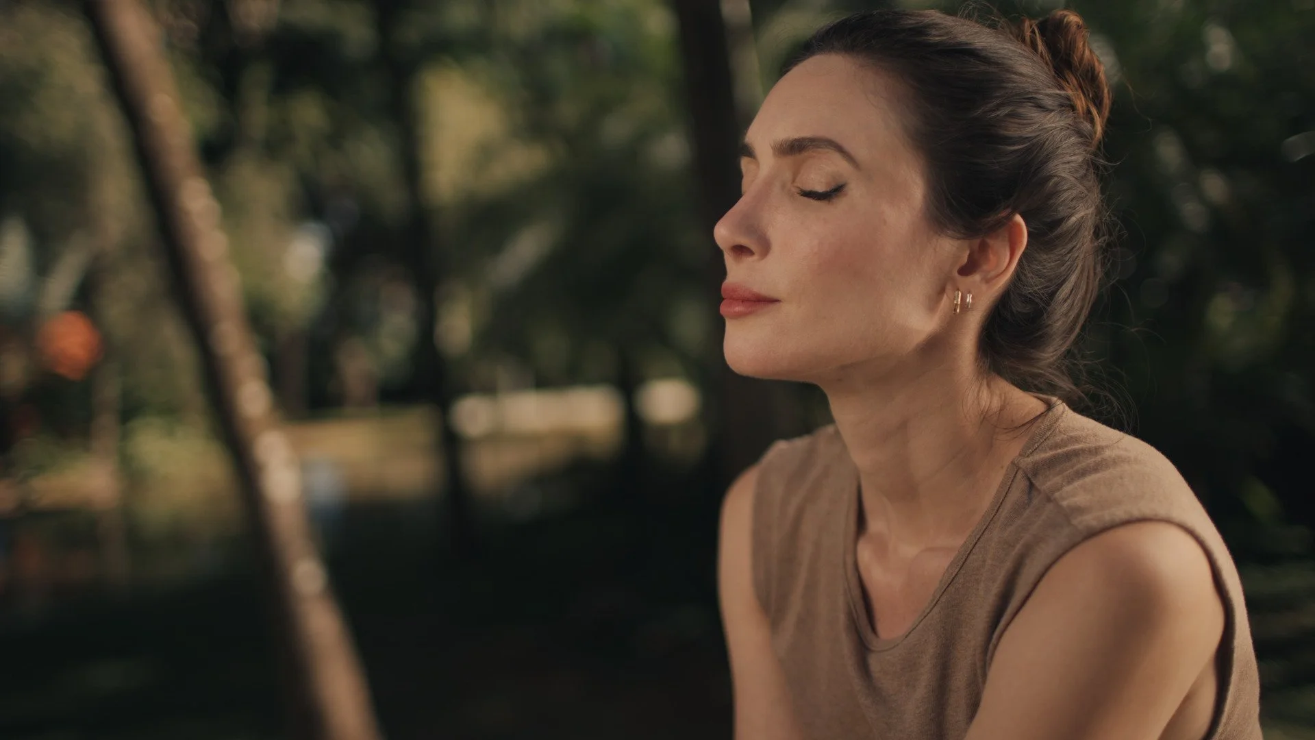 Retrato de uma mulher com expressão serena e olhos fechados, ao ar livre, com fundo de árvores e luz natural.