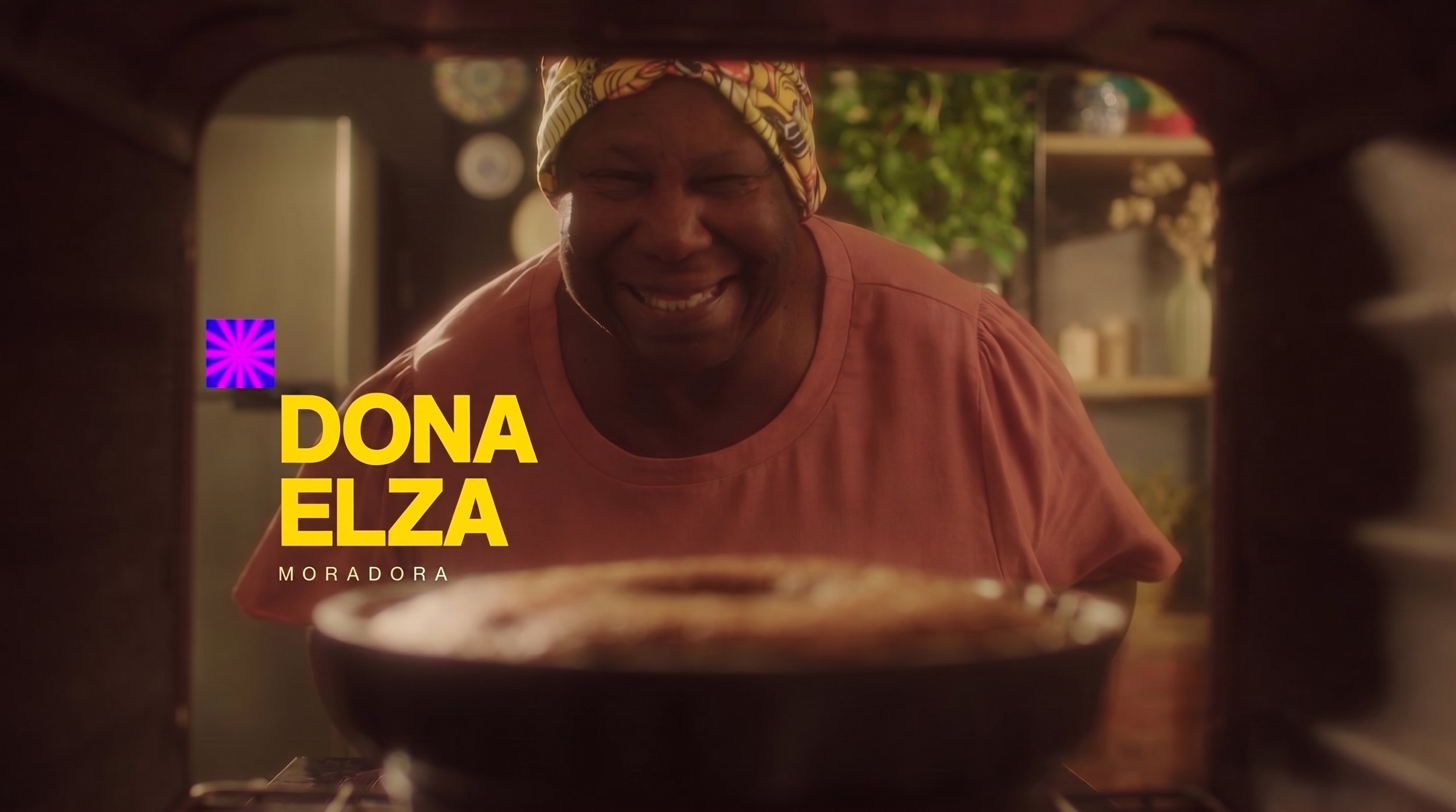 Cena de uma mulher sorridente assistindo um forno ou micro-ondas de dentro. Ela usa um turbante colorido e uma blusa de cor salmão. O ambiente é aconchegante, com decoração na parede e plantas ao fundo. Texto na imagem diz: Dona Elza, Moradora.