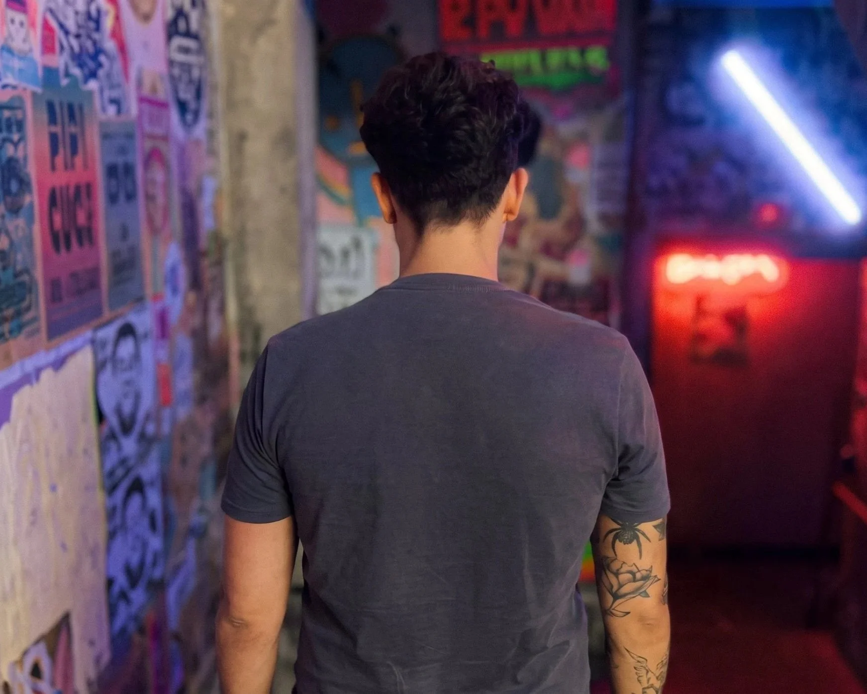 Pessoa com cabelo cacheado escuro, visto de costas, usando uma camisa cinza escura, com tatuagens no braço direito, em um ambiente com murais e letreiros coloridos ao fundo.