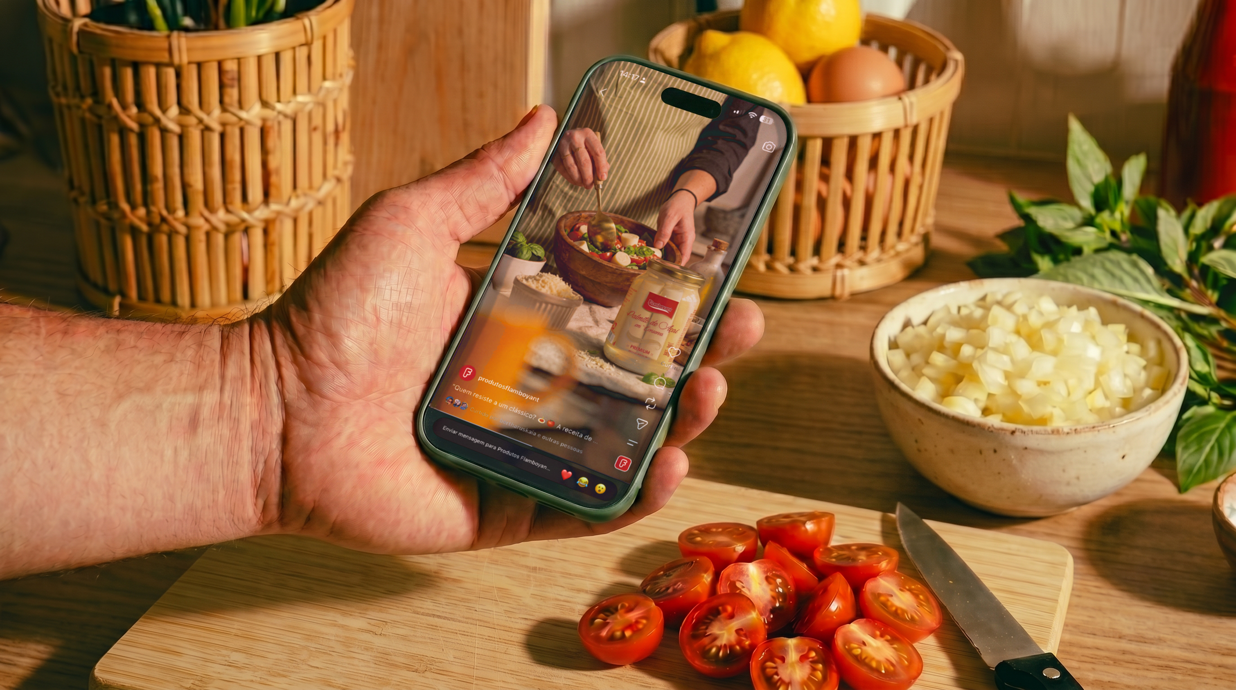 Pessoa segurando um celular enquanto fotografa ingredientes para uma salada, incluindo tomates e queijo em uma bancada de cozinha, com cestos de frutas ao fundo.