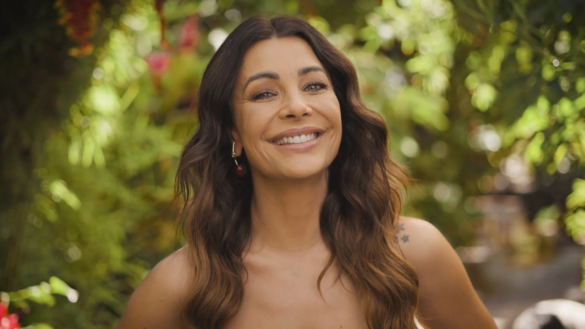 Mulher sorridente com longos cabelos castanhos ondulados, usando brincos, ao ar livre com fundo de folhagem verde.