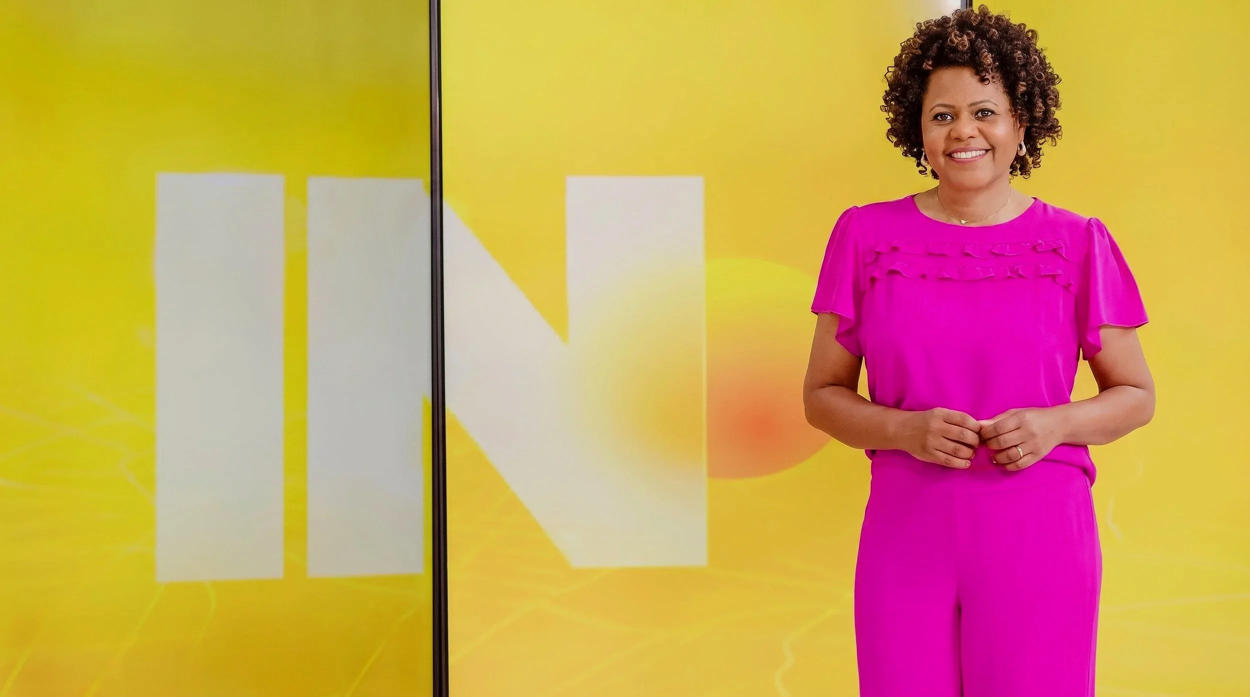 Mulher sorridente com cabelo cacheado usando vestido rosa em frente a fundo amarelo com formas geométricas.