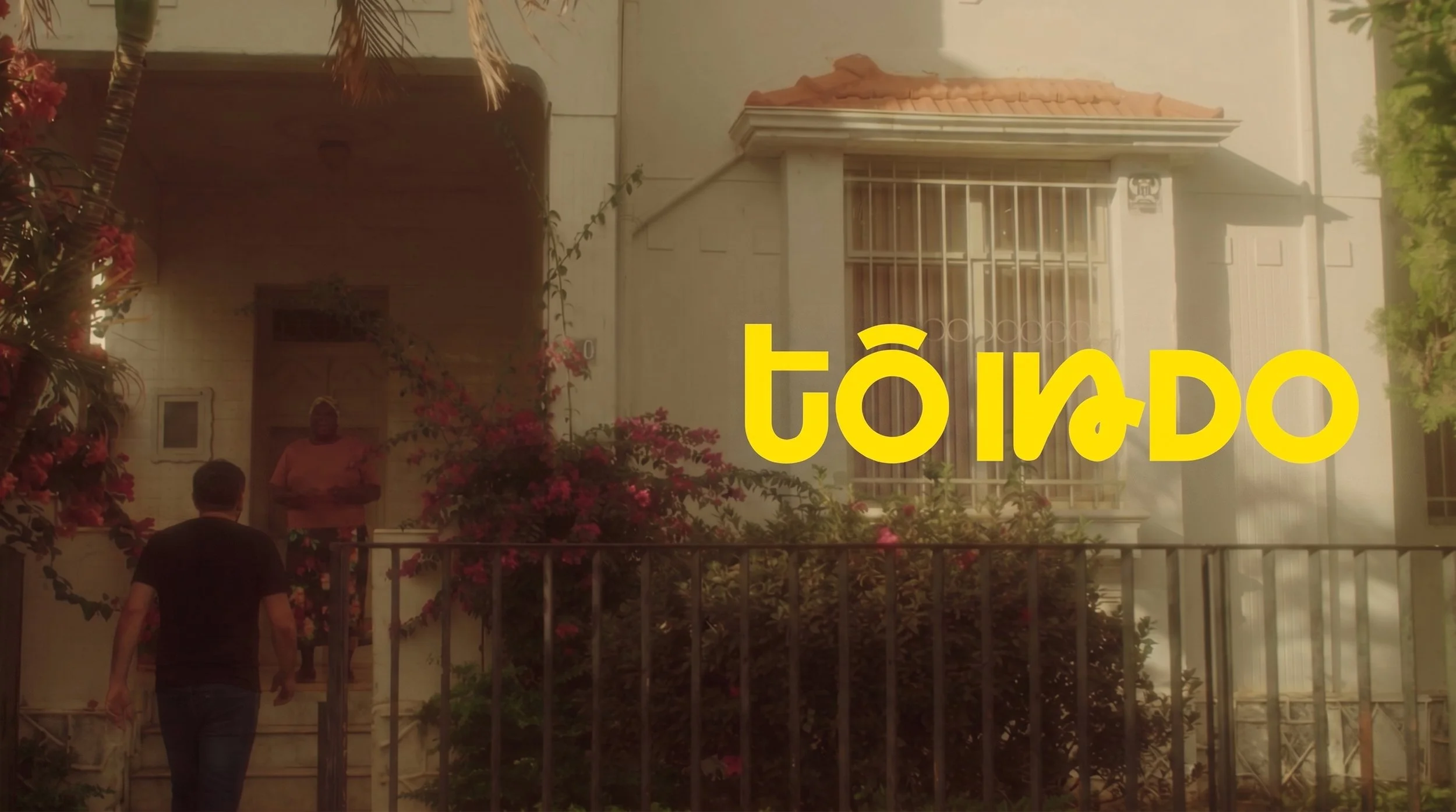 Imagem de uma entrada de casa com uma grade de ferro na porta e flores ao redor, com uma pessoa de costas caminhando em direção à porta. Logotipo em destaque amarelo com a palavra "tô indo".