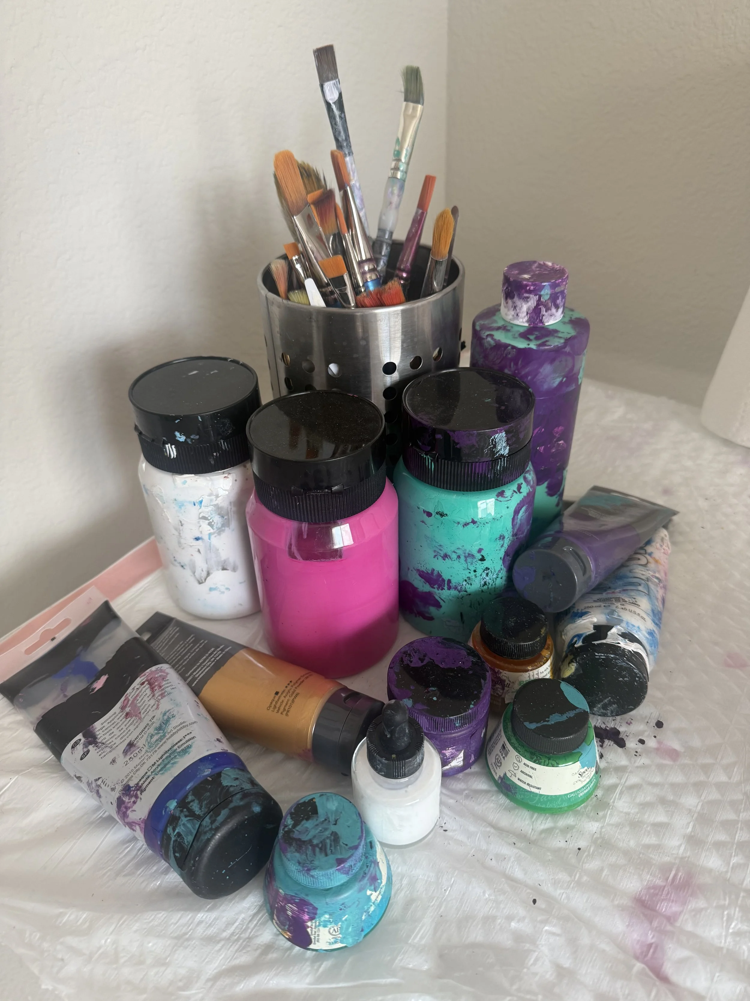 A collection of paint bottles and brushes on a table, with paint splatters and some overlapping.