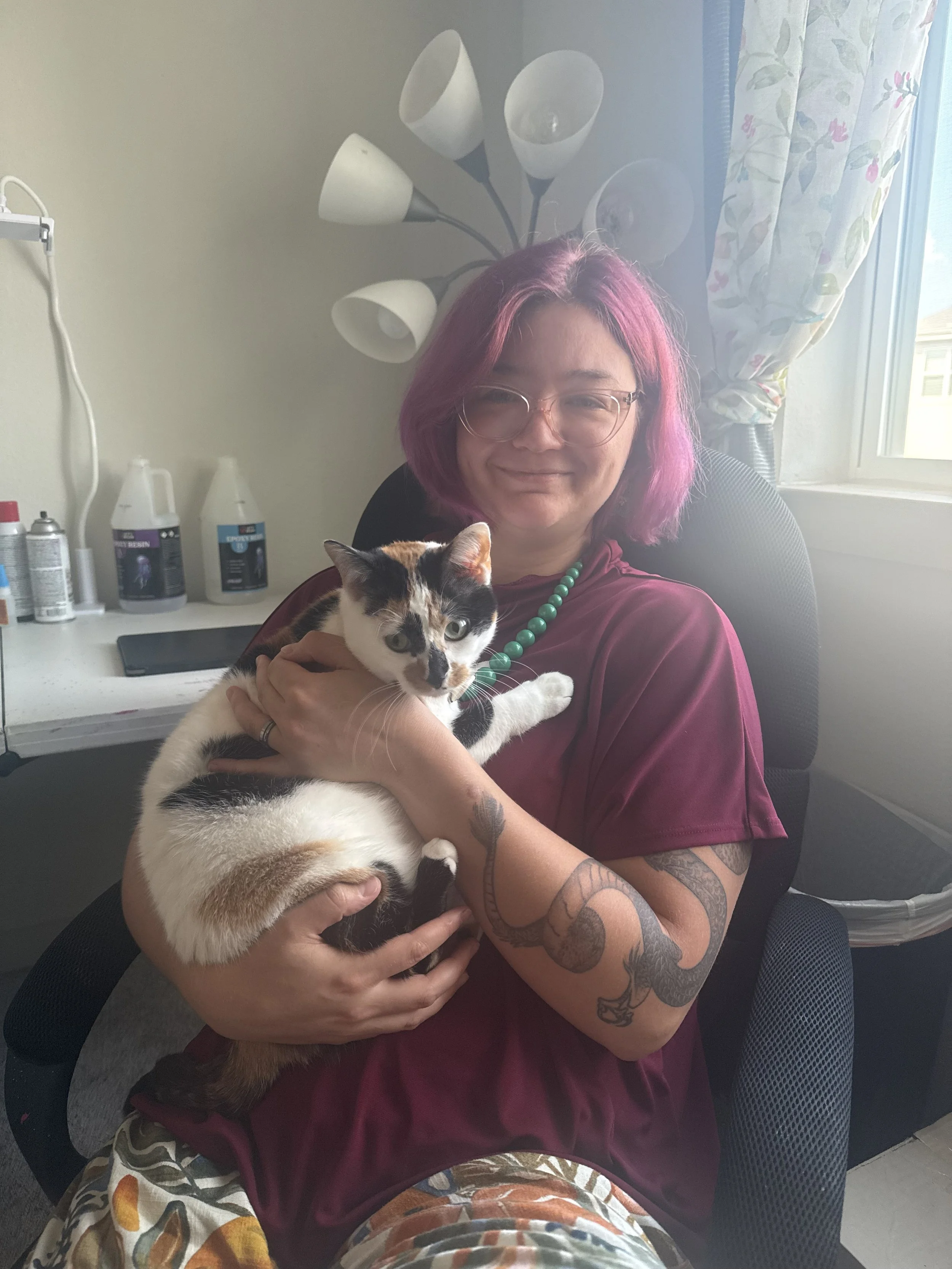 A woman with purple hair, glasses, and tattoos holding a calico kitten in a room with a window, a floor lamp, and bottles on a desk.