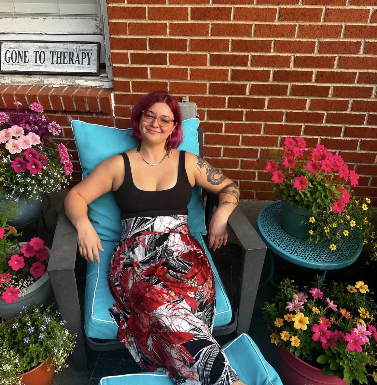 A woman with pink hair and glasses sitting comfortably in a chair on a patio, surrounded by colorful flowers, with a brick wall behind her and a sign that reads 'Gone to Therapy' in the background.
