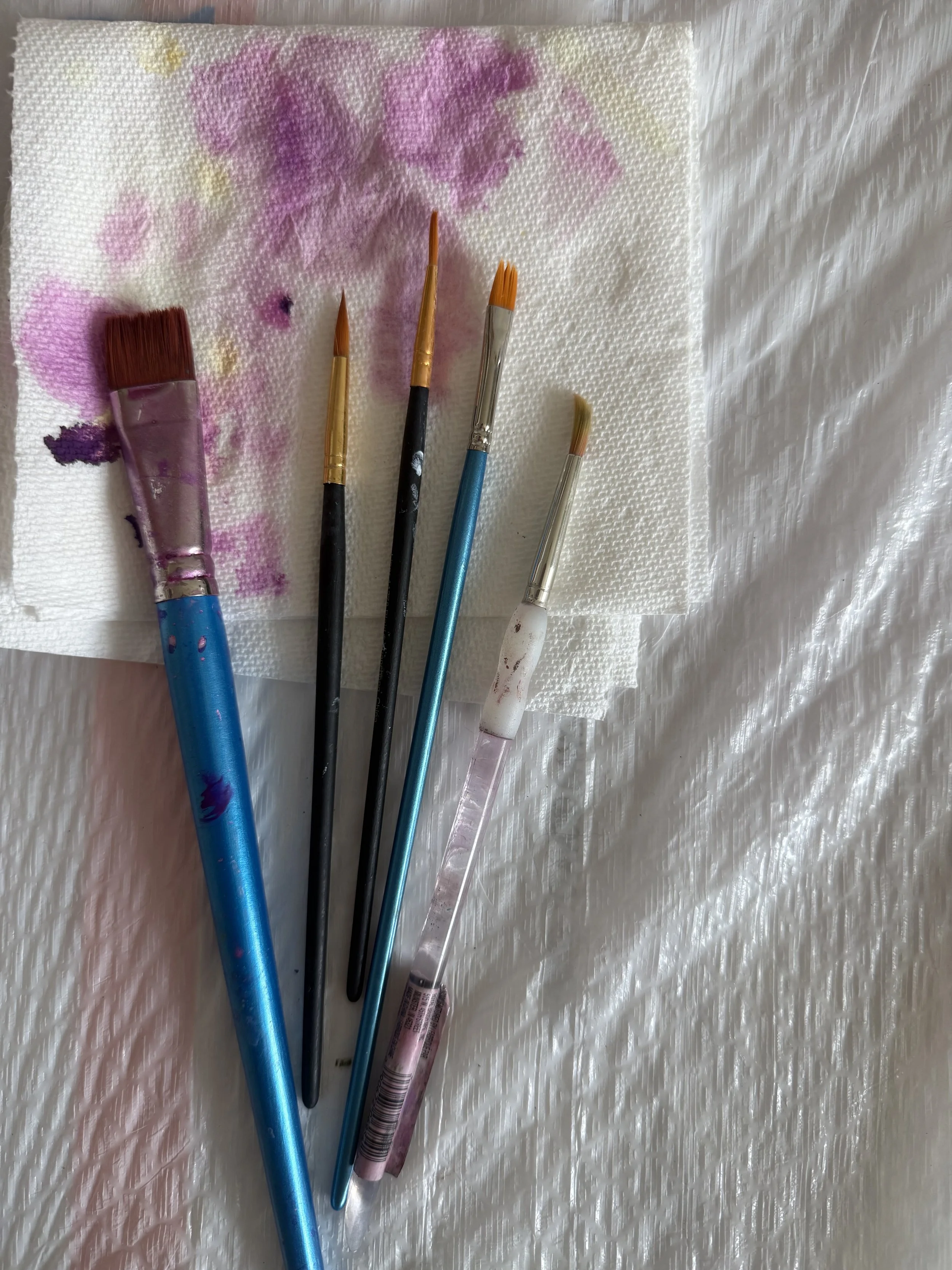 Several paintbrushes of various sizes and a pen resting on a paper towel with splashes of pink, purple, and white paint, on a textured surface.