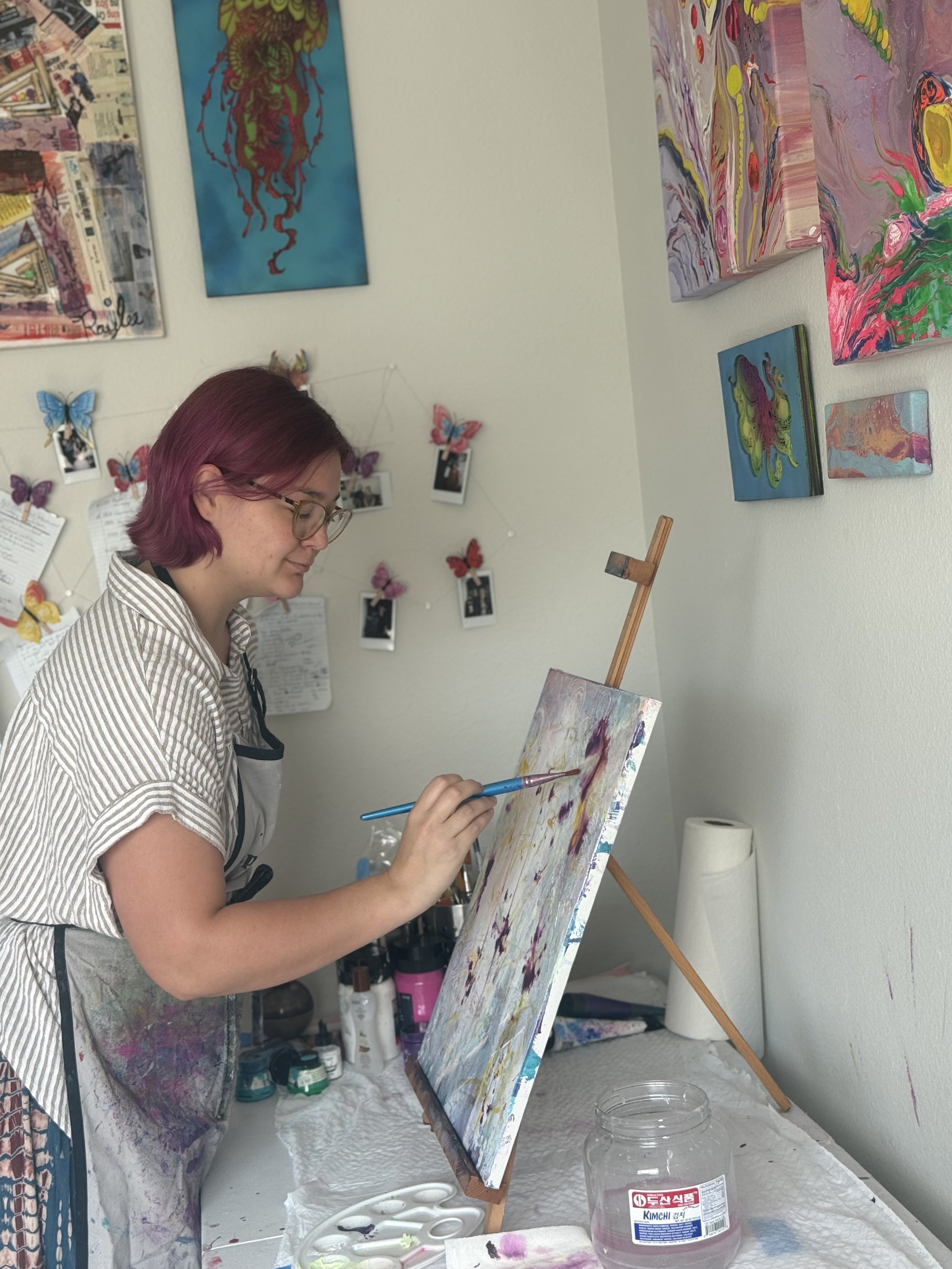 A woman with purple hair and glasses painting on a canvas in a room decorated with colorful abstract paintings and butterfly photos. She is wearing a striped shirt and a paint-stained apron, holding a paintbrush while working on her artwork.