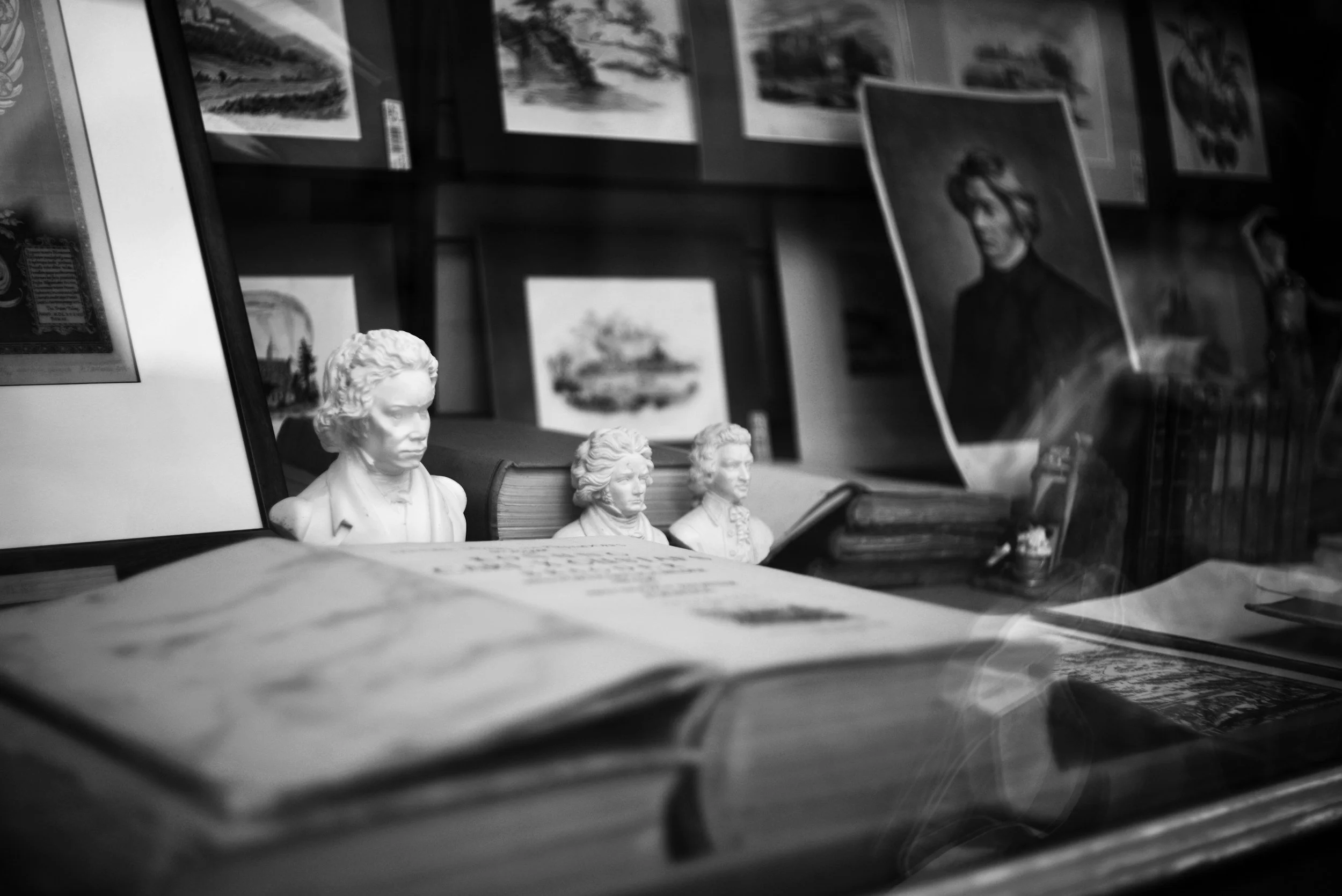 A black-and-white photograph of a shelf displaying portraits and busts of famous historical figures, with books and papers in the foreground.