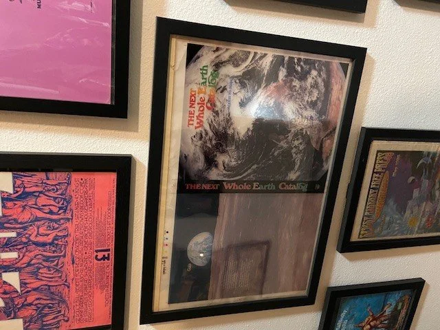 Framed poster titled "The Next Whole Earth Catalog" featuring an image of Earth from space, mounted on a wall among other framed posters.