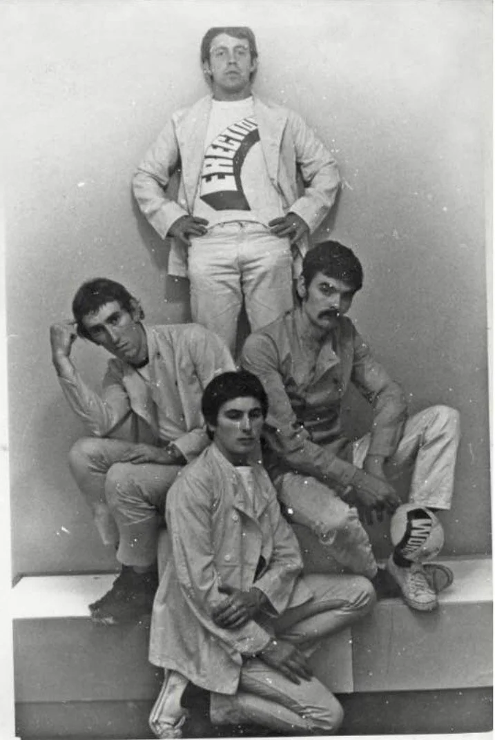 Black and white photo of four young men posing against a plain wall, with one standing and three sitting or crouching on a ledge. The standing man is wearing a jacket and t-shirt, with hands in pockets. The sitting men wear casual clothing, with one resting his head on his hand, another sitting with crossed legs, and the third sitting on the ledge with a soccer ball nearby.