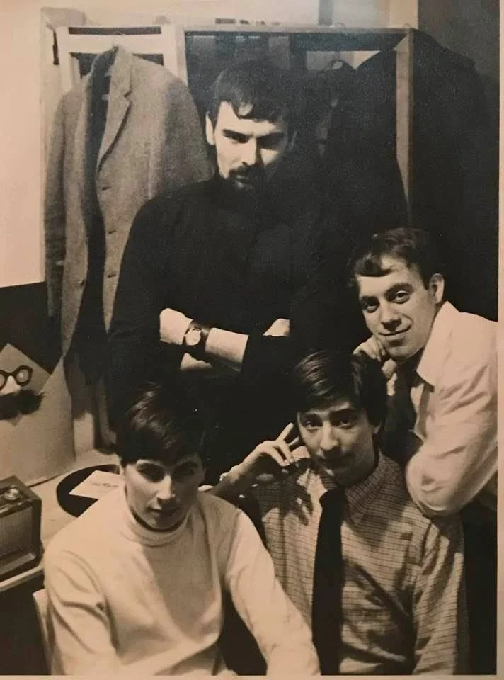 A black and white photo of four young men posing together indoors, with one standing in the background and three sitting in the foreground. The person in the background has crossed arms and is wearing a dark shirt. The three in the foreground are casually posed, with one resting his hand on his head and another making a peace sign near his temple, all smiling or with relaxed expressions.