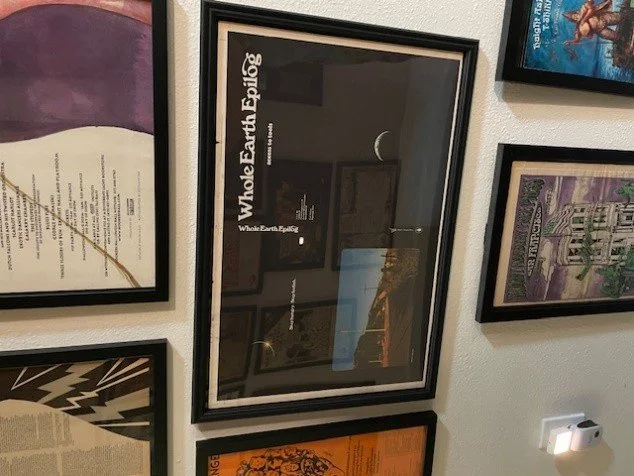 Framed poster titled 'Whole Earth Epilog' hanging on a white wall surrounded by other framed posters.