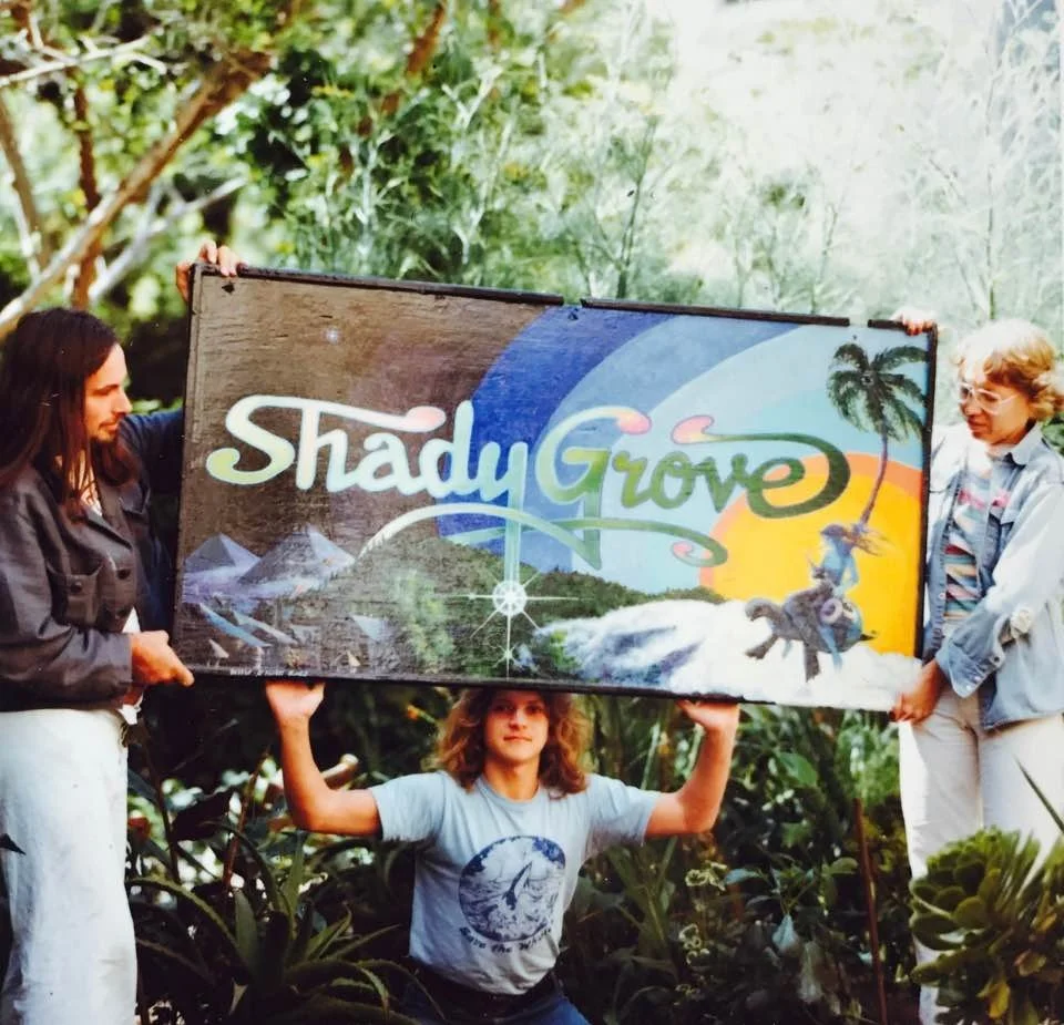 Three people holding a colorful sign that reads 'Shady Grove' in a lush, green outdoor setting.