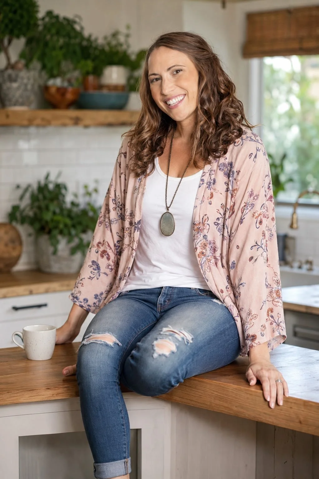 A woman with curly brown hair smiling and sitting on a kitchen counter, wearing a white t-shirt, a floral pink kimono, ripped jeans, and a long pendant necklace.