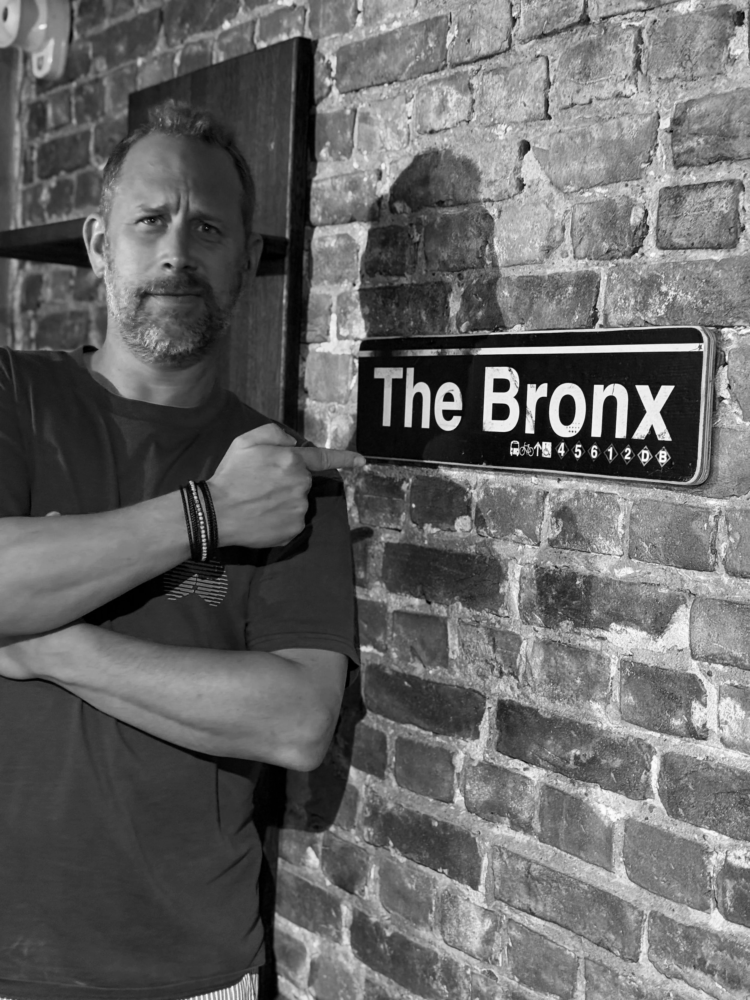 A man with a beard and short hair pointing to a sign that reads 'The Bronx' on a brick wall. The man is wearing a dark shirt and wristbands.