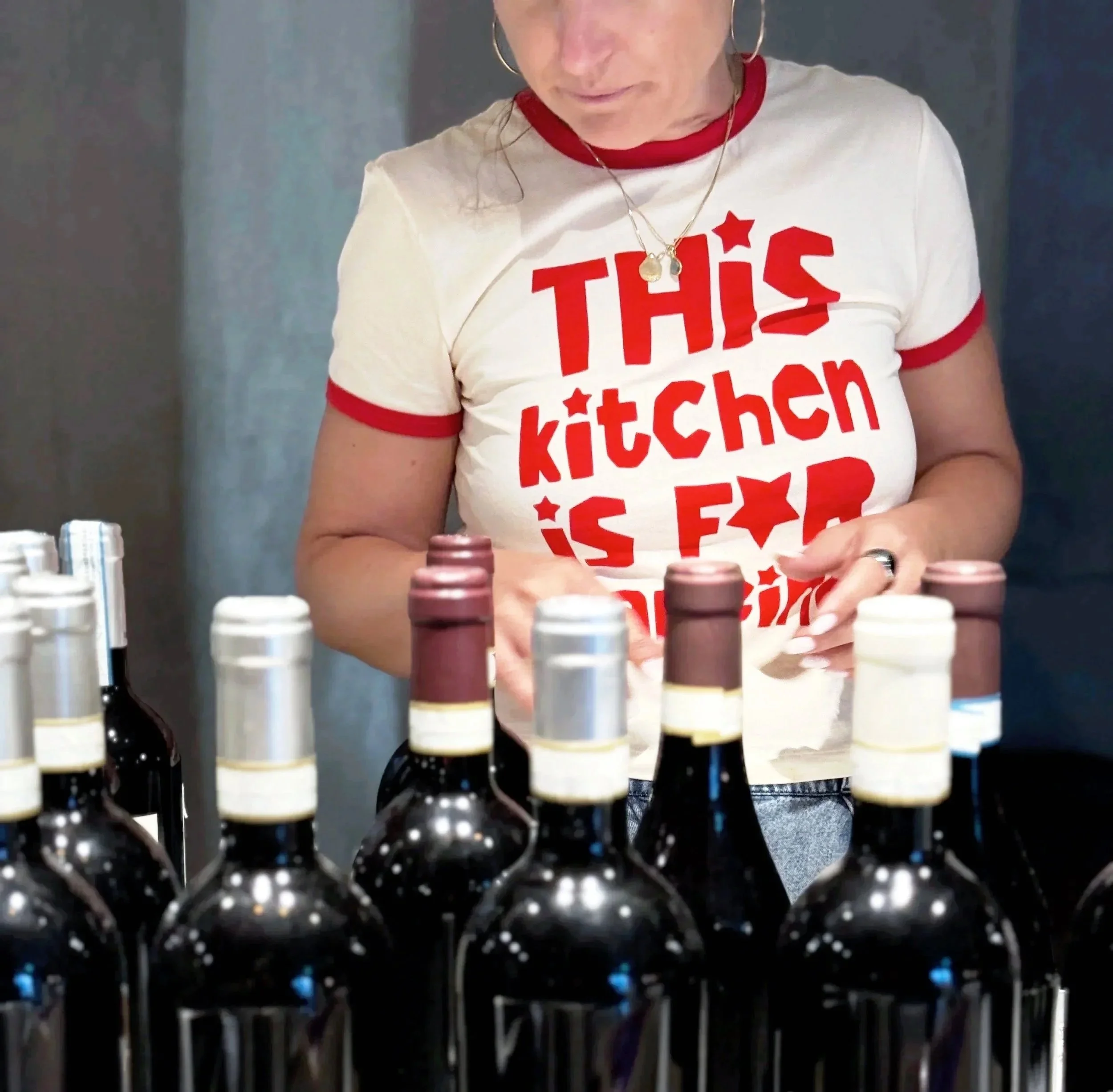 A woman wearing a white T-shirt with red text is working at a table with various bottles, possibly nail polish or other small cosmetic bottles, in front of her. The woman has hoop earrings and a necklace with pendants.