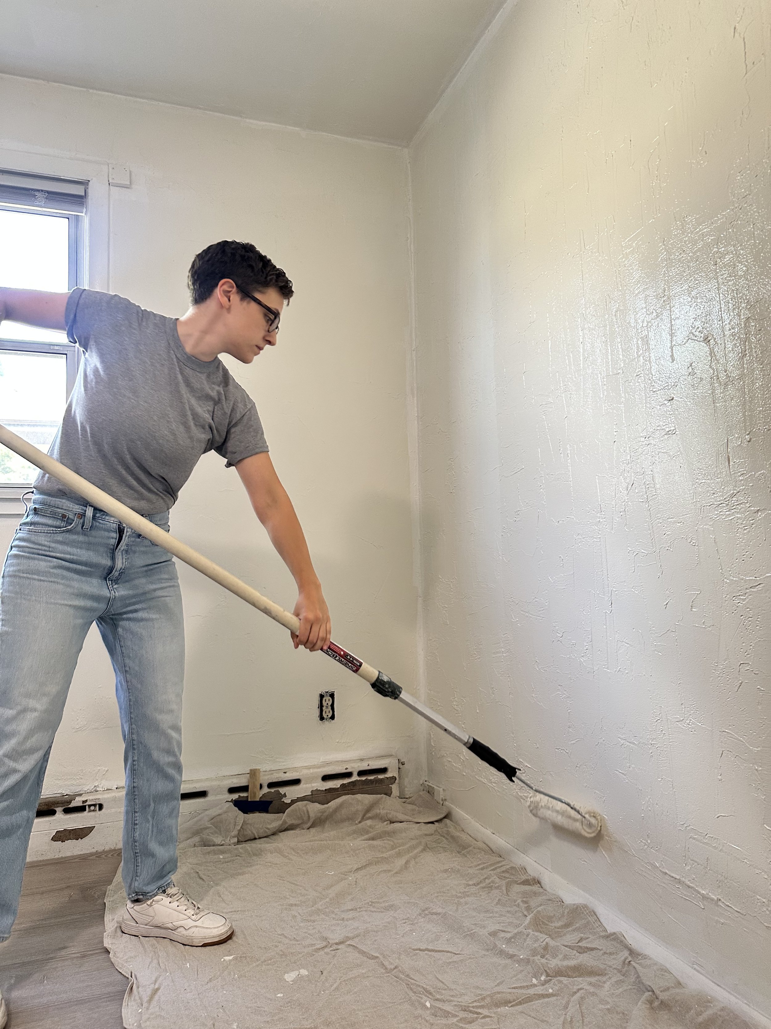 A person wearing glasses, a gray t-shirt, light blue jeans, and white sneakers is painting a wall with white paint using a roller brush in a room with a window and a vent on the floor.