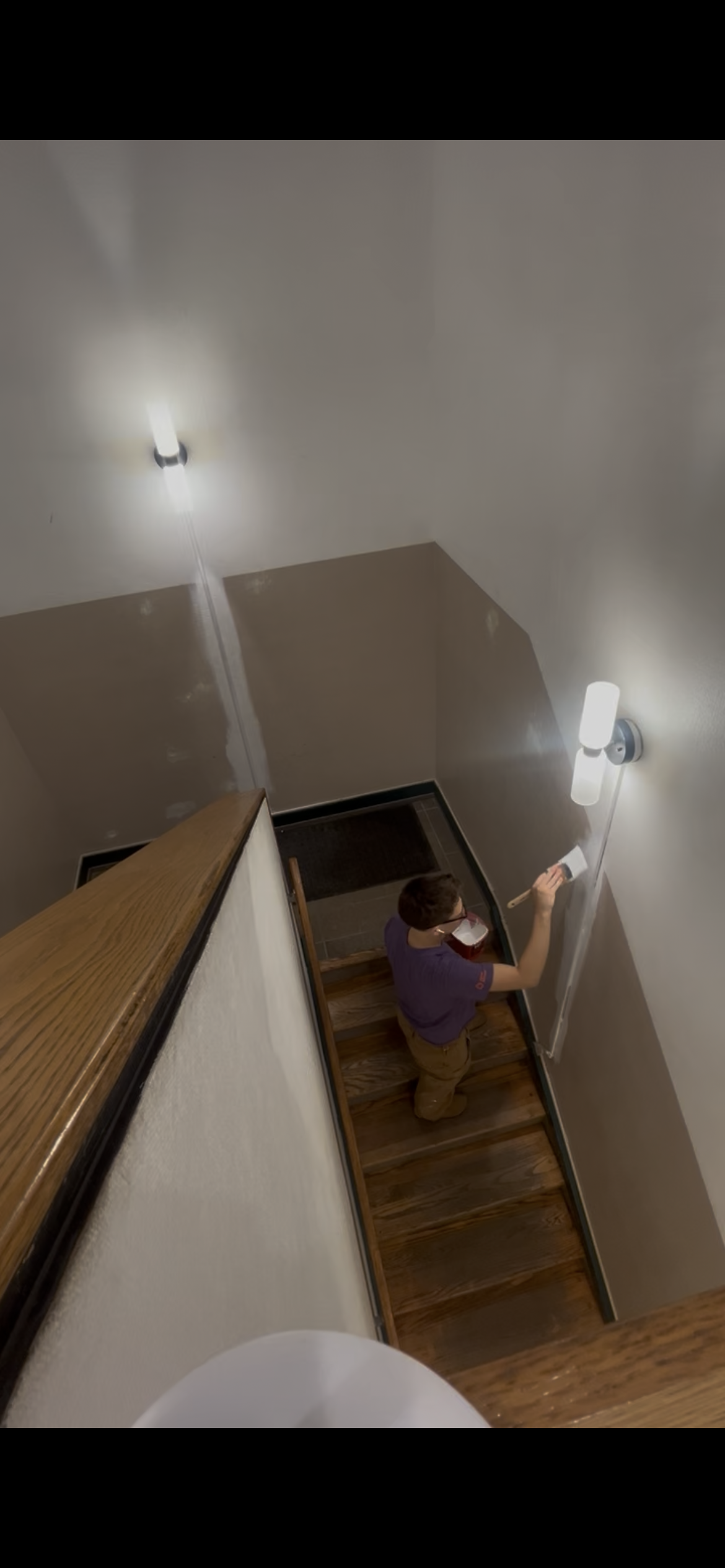 Person painting the wall in a staircase area with a paintbrush and a bucket, viewed from the top of the stairs.