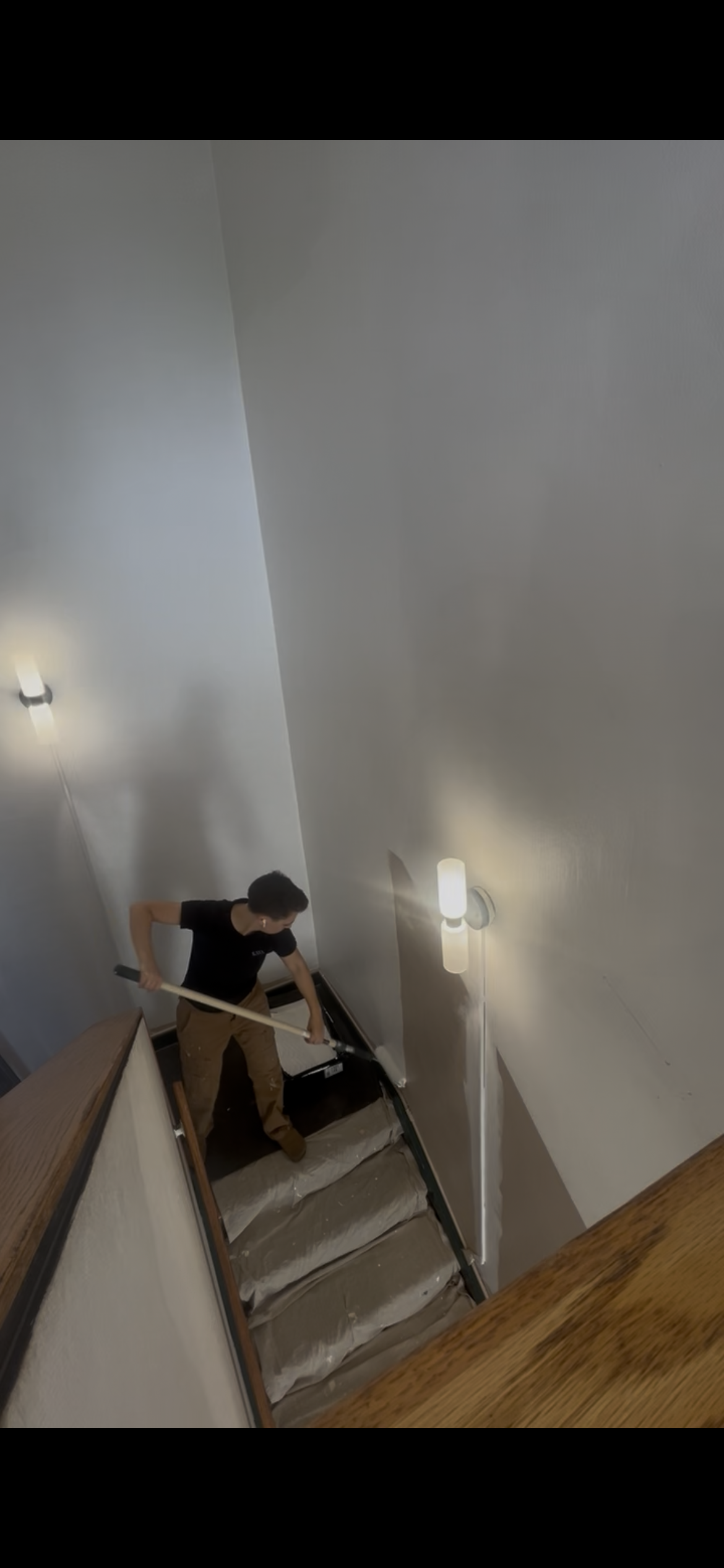 A person is cleaning or painting the wall of a staircase hallway with a long-handled tool. The staircase has a wooden handrail and carpeted steps, and is illuminated by wall-mounted light fixtures.