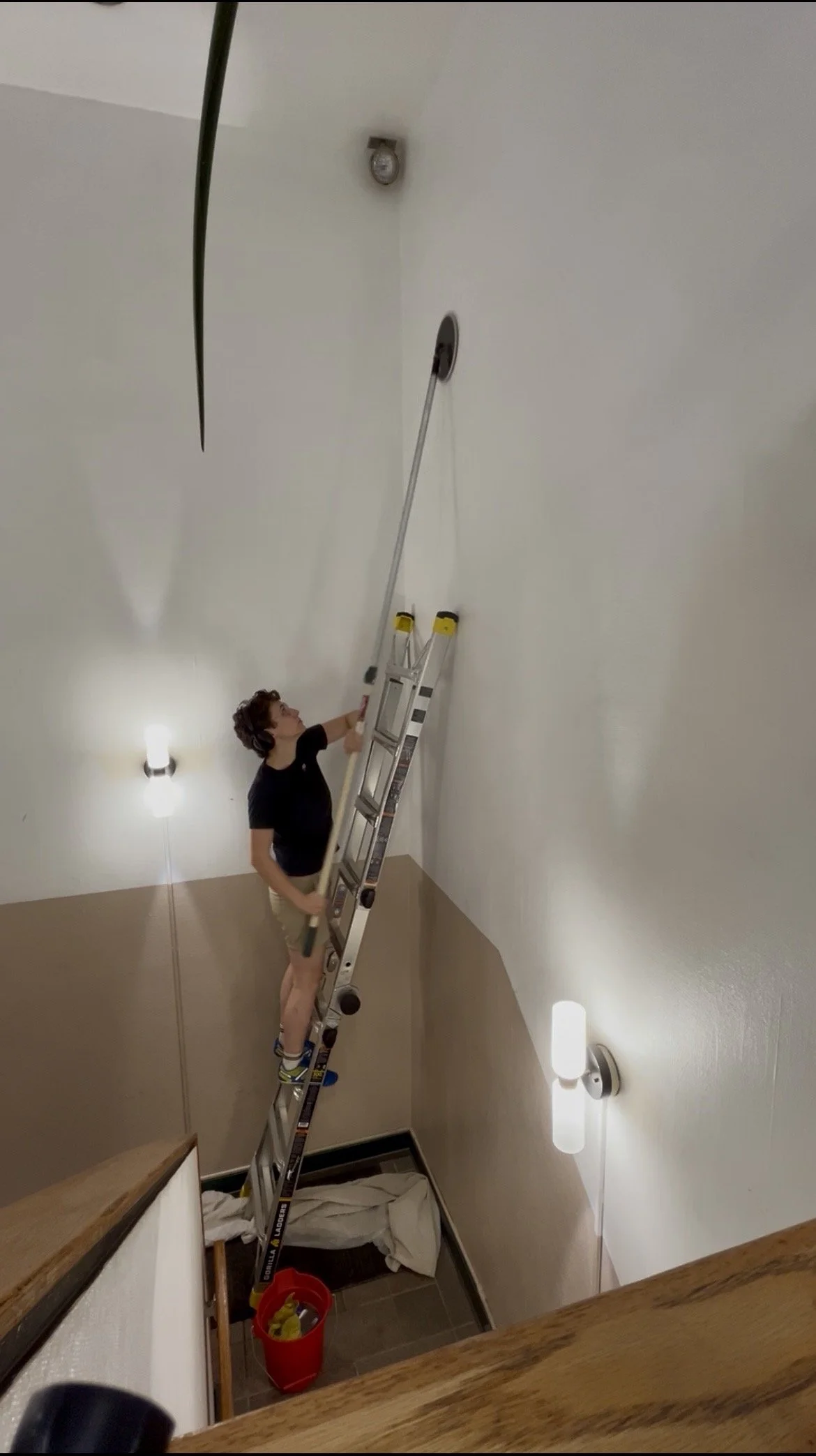 A person stands on a ladder, reaching up to change a ceiling light fixture in a room. The room has tan walls, wall-mounted lights, and a shower with a handheld showerhead. There is a red bucket and a blanket on the floor.