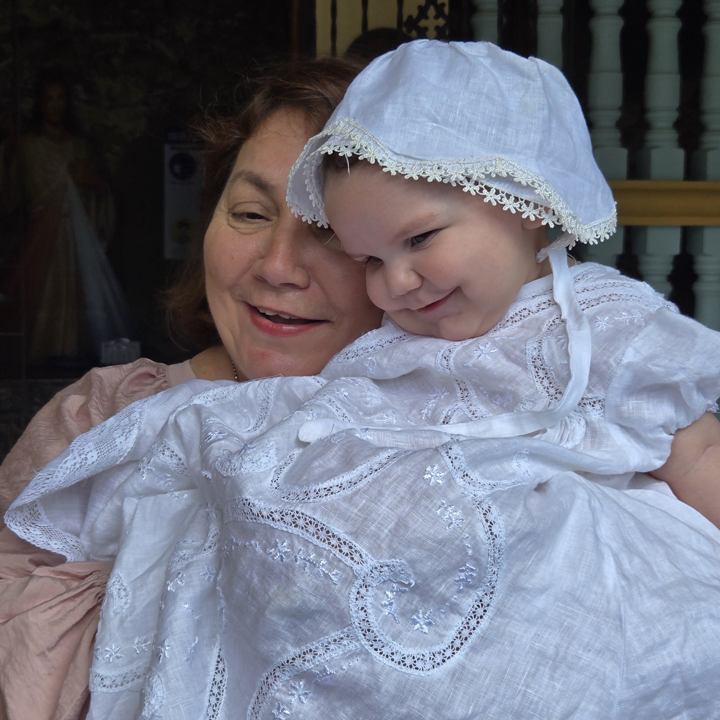 Close-up of an elderly woman with brown hair holding a young girl wearing a white lace dress and matching bonnet, both smiling, in a warm and joyful moment.