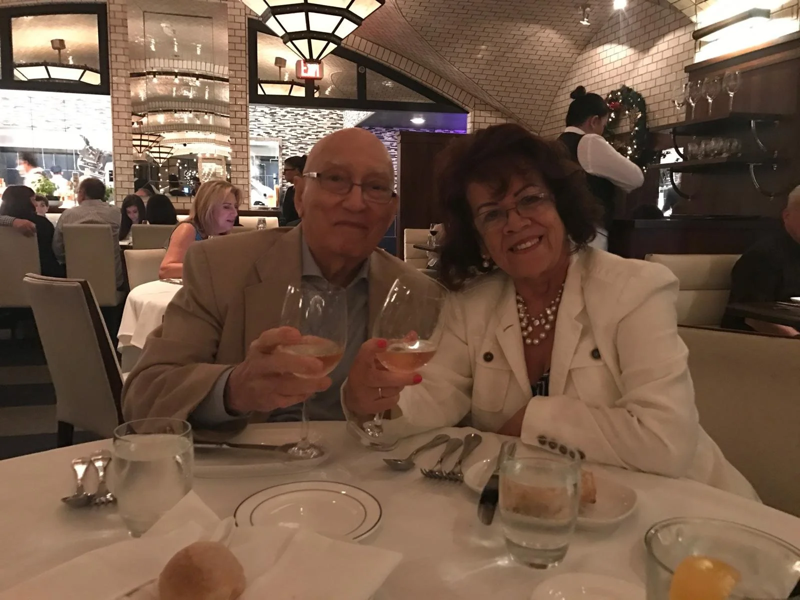 A senior man and woman sitting at a restaurant table, holding wine glasses, with other diners and a waiter in the background.