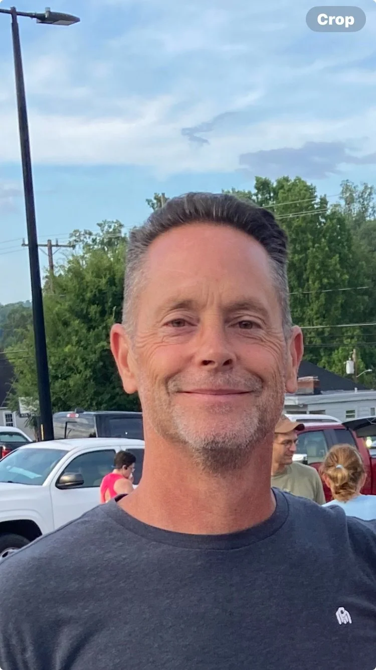 A smiling man with short hair and a beard, wearing a dark T-shirt, standing outdoors in a parking lot with several cars and people in the background, under a partly cloudy sky.