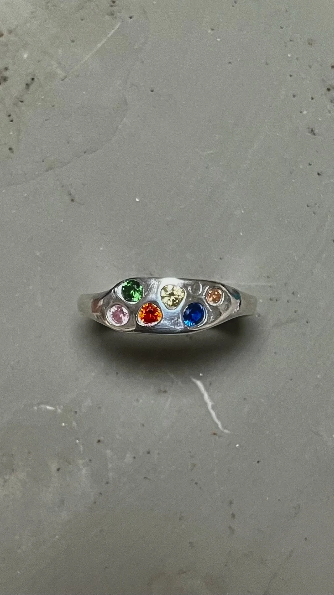 A silver ring with seven colorful gemstones: green, yellow, orange, pink, red, blue, and peach, on a gray surface.
