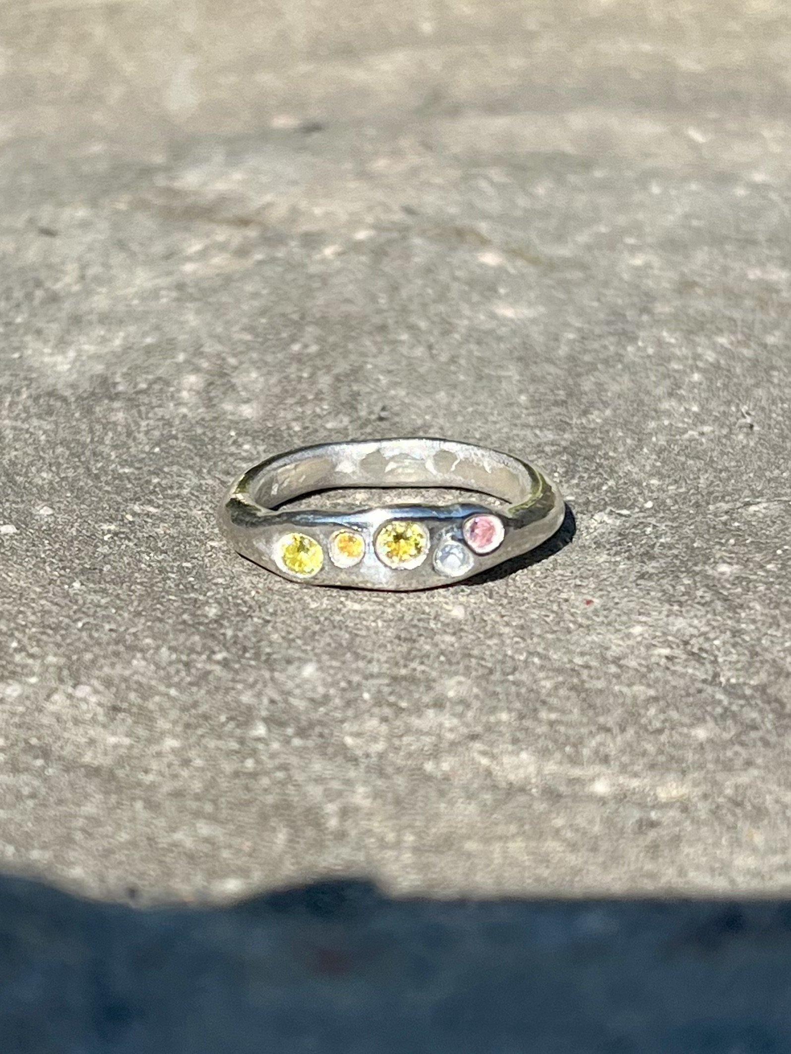 A silver jewelry ring with multiple colored gemstones on a gray textured surface.