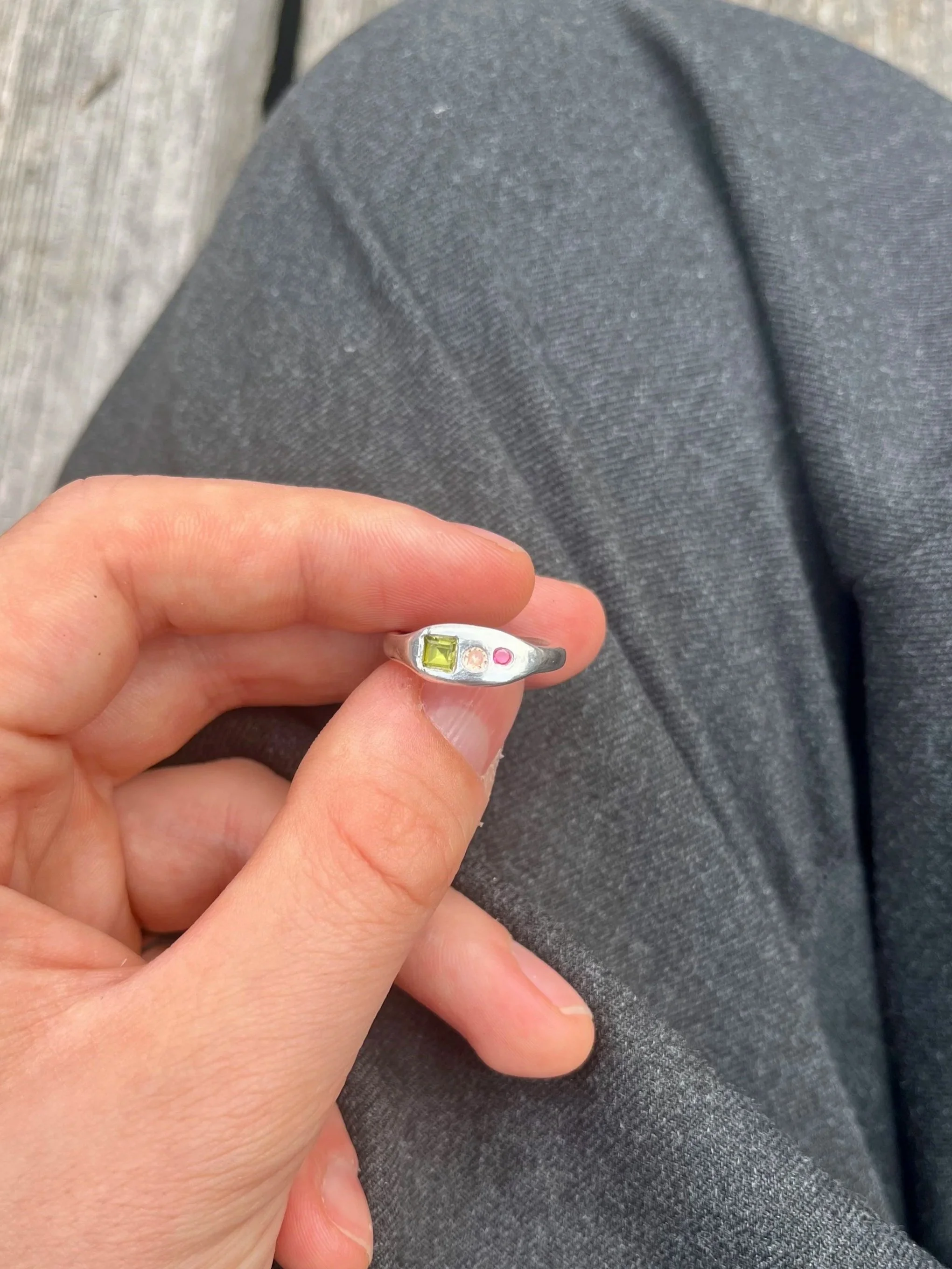 A person holding a silver ring with three gemstones: one yellow, one pink, and one clear, set in a row.