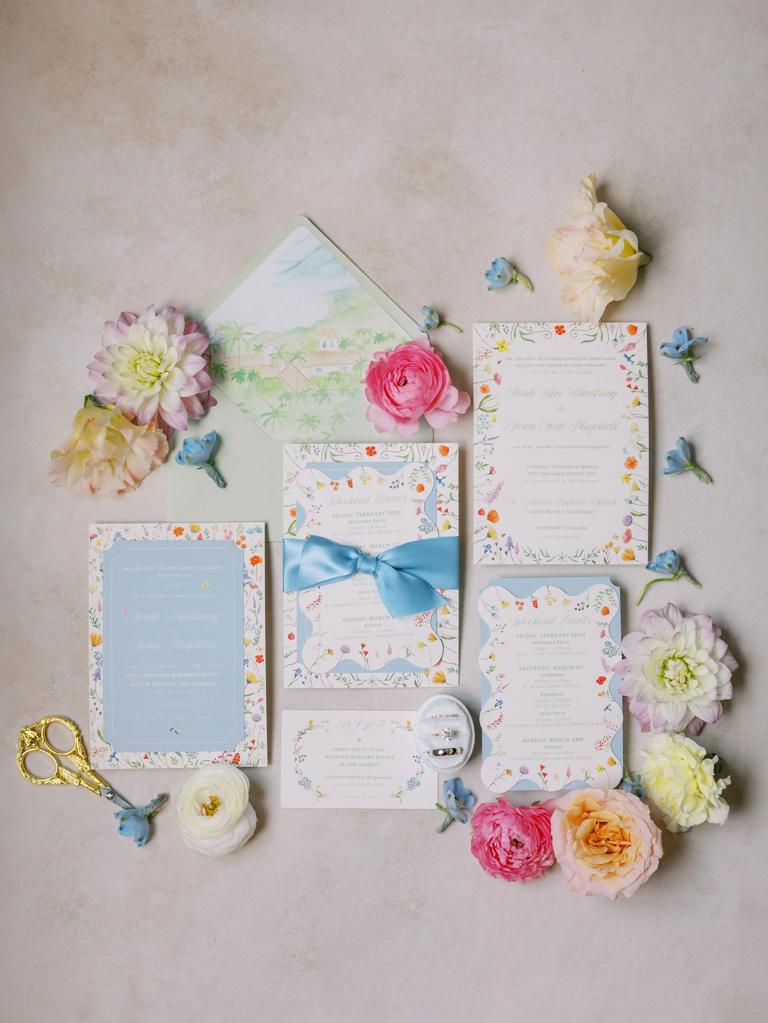 Wedding invitation and RSVP cards surrounded by colorful flowers and small decorative scissors on a light background.