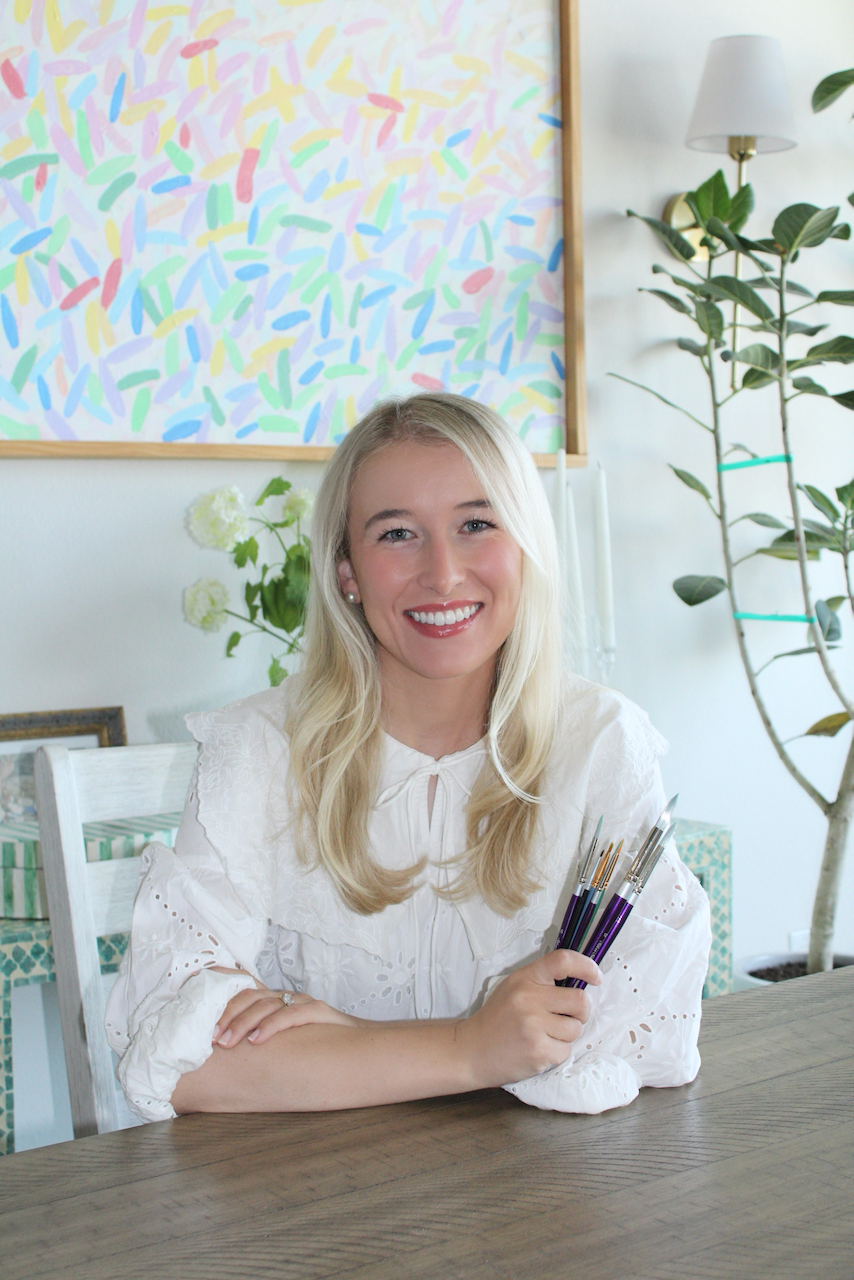 A blonde woman smiling and sitting at a wooden table, holding paintbrushes, with a colorful abstract painting, green plants, and a white wall with a lamp in the background.